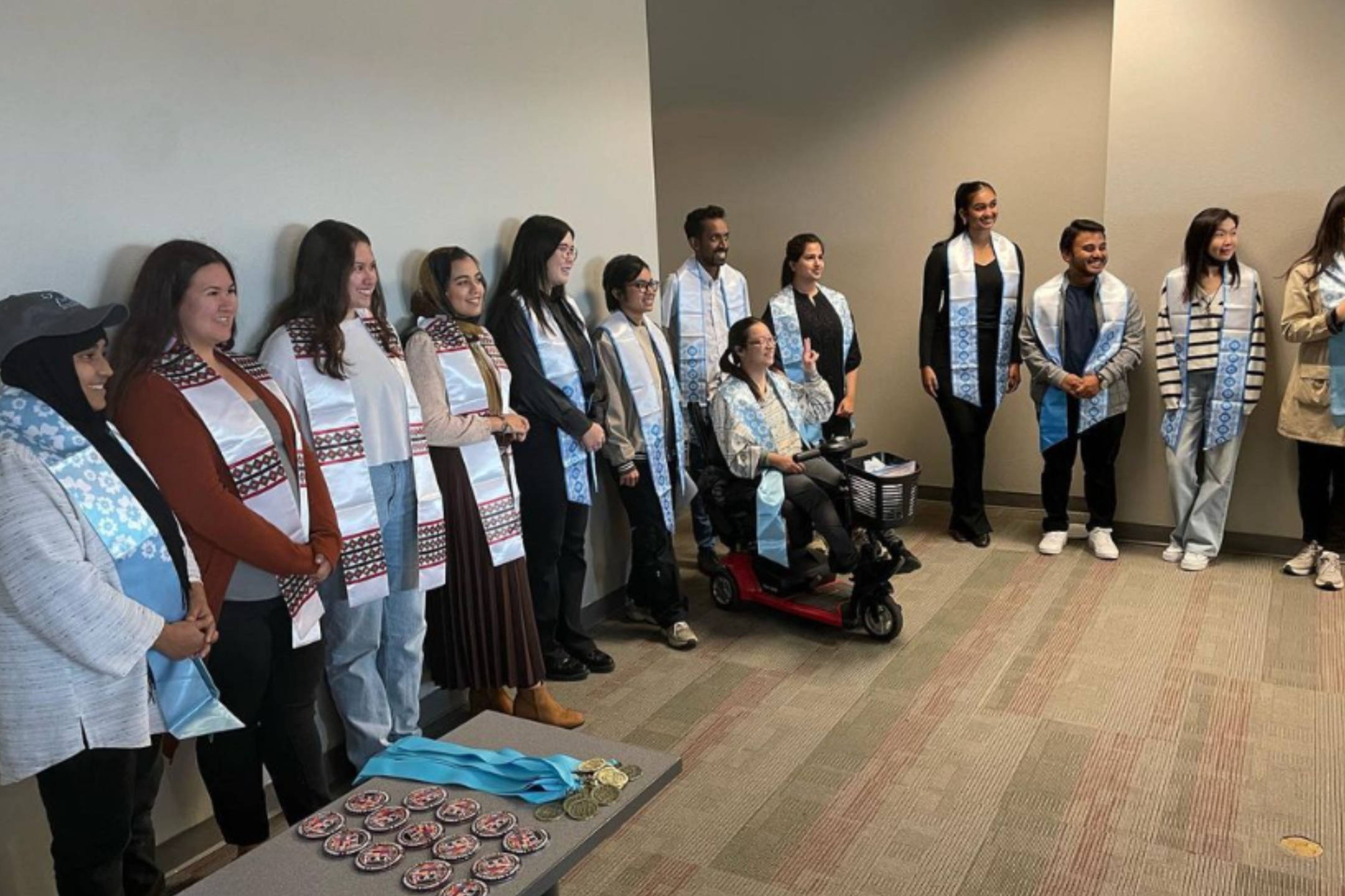 The image shows a group of diverse people posing together in what appears to be a celebratory or academic context. The group stands against a plain wall in a room with a beige carpeted floor. There are thirteen individuals in the group, including a person in a red mobility scooter. Most of the people are wearing white sashes with intricate geometric patterns over their shoulders. Some of the sashes have blue and white floral designs, while others feature red, black, and white patterns. The individuals are dressed in various casual to semi-formal outfits, including shirts, blouses, sweaters, and pants. On a table in the foreground, there are several circular objects, possibly medals or tokens, with attached blue ribbons and intricate designs.