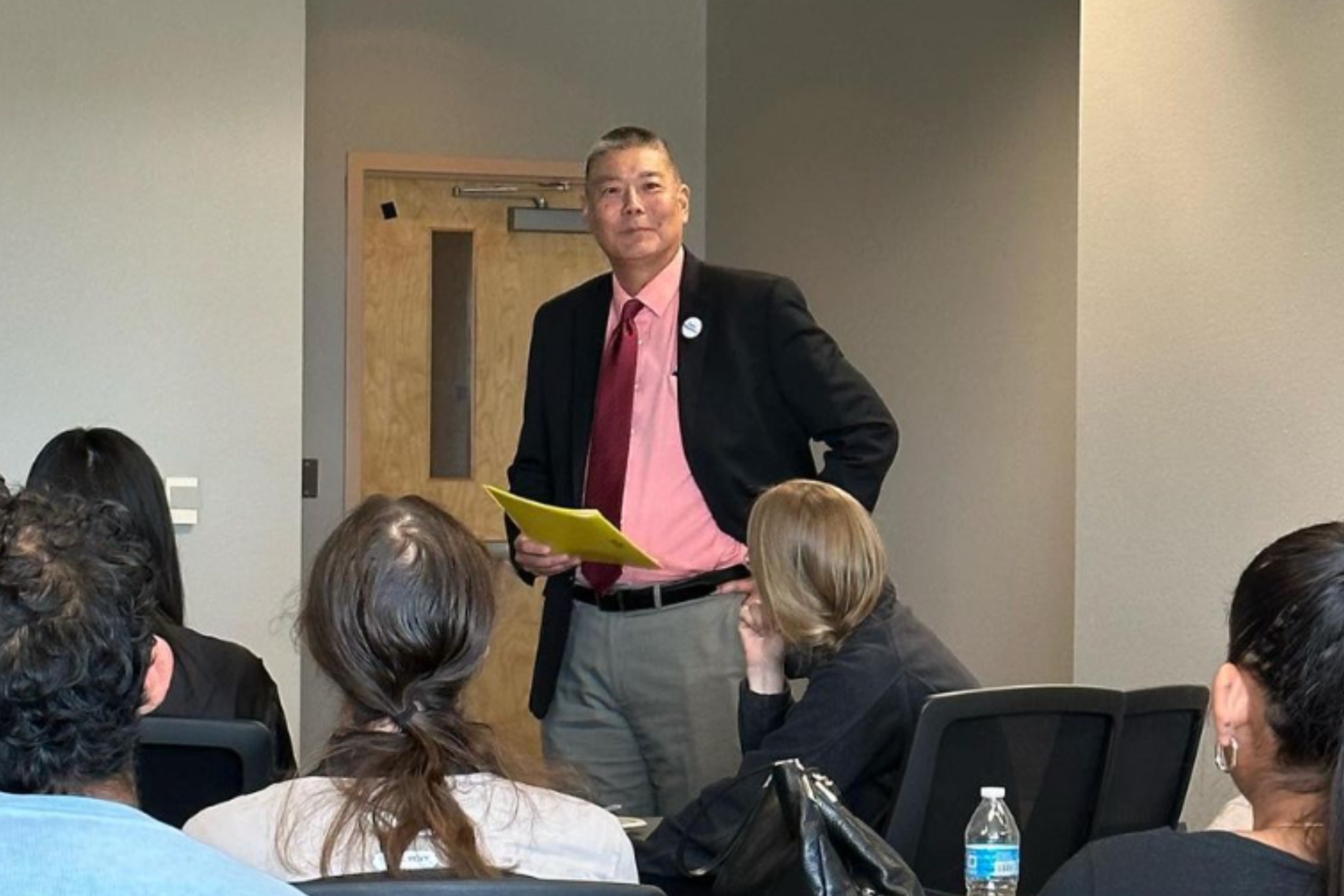  The image depicts a man standing in front of a room of seated individuals, presumably giving a presentation or lecture. The man is wearing a black blazer, a pink dress shirt, a red tie, and gray trousers. He holds a yellow folder in his left hand and has his right hand resting on his hip. The audience members are captured from behind; their heads and shoulders are visible but their faces are not clearly seen. In front of the audience, a clear plastic water bottle is noticeable on a table. The background features a plain wall with neutral color tones and a closed light wooden door with a small rectangular window at the top.