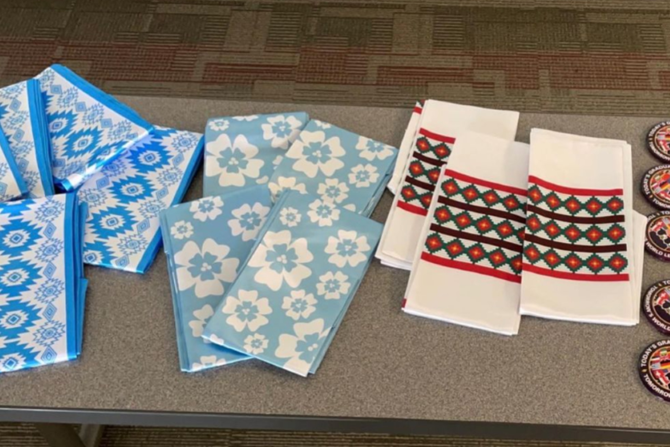 The image depicts a display of folded cloth items, likely napkins or handkerchiefs, arranged on a grey tabletop. The cloths are presented in three distinct patterns and colors. On the left, there are blue napkins with geometric patterns in white and various shades of blue. In the middle, the napkins are light blue with a repeating white floral pattern. On the right, there are white napkins with intricate black, red, and green geometric patterns, bordered with red stripes. Also visible to the far right are several round patches with multicolored designs, though the details are not clear enough to be described here. The background consists of a different pattern in shades of brown, grey, and red, likely a carpet.