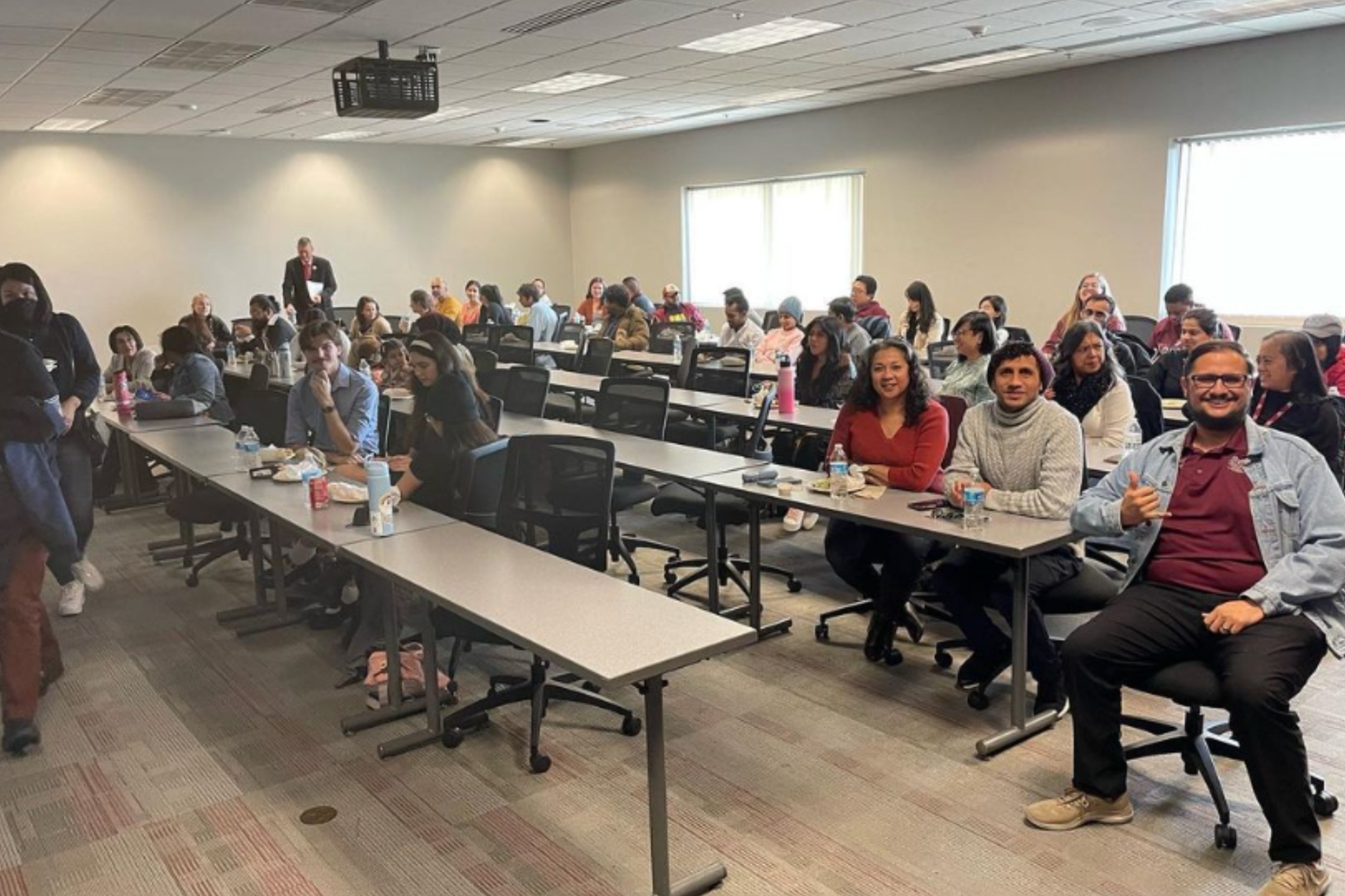 The image depicts a large, well-lit classroom filled with over 30 people seated at long tables arranged in rows. The room's walls are light gray, and the floor is carpeted with a pattern of neutral tones. At the front, a male figure stands near a wall, appearing to read from some papers. The attendees, of diverse backgrounds, are engaged in conversations or looking towards the camera, with some smiling. The tables are cluttered with various items, including notebooks, bottles, and snacks. Above the room, a ceiling-mounted projector is visible. Large windows on the right side of the room allow natural light to enter, contributing to the bright atmosphere.