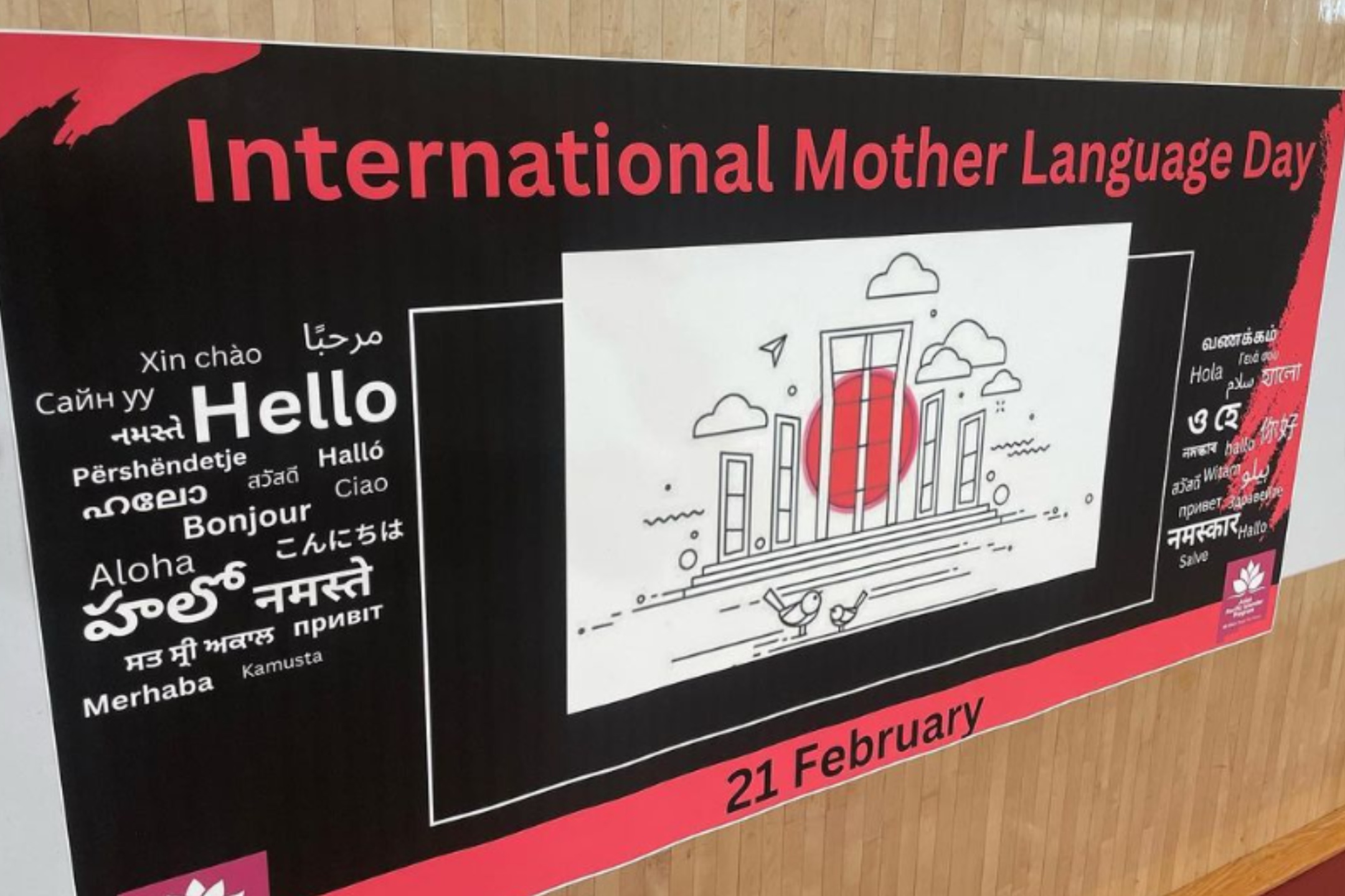 The image is a horizontal banner hung against a light wooden wall. The banner primarily has a black background with red and white accents. At the center of the banner is an illustration featuring a series of stylized buildings and a sun or circle in red, with small clouds and birds around it in black lines.  Above the illustration, large red text reads "International Mother Language Day." Surrounding the illustration on both sides are the words "Hello" in various languages and scripts including English, Arabic, Chinese, Hindi, Punjabi, Russian, and many others in both white and red text.  The bottom of the banner, which is red, contains the date "21 February" in bold black text at the center. In the bottom left and right corners of the banner, there are logos with white symbols on red backgrounds.