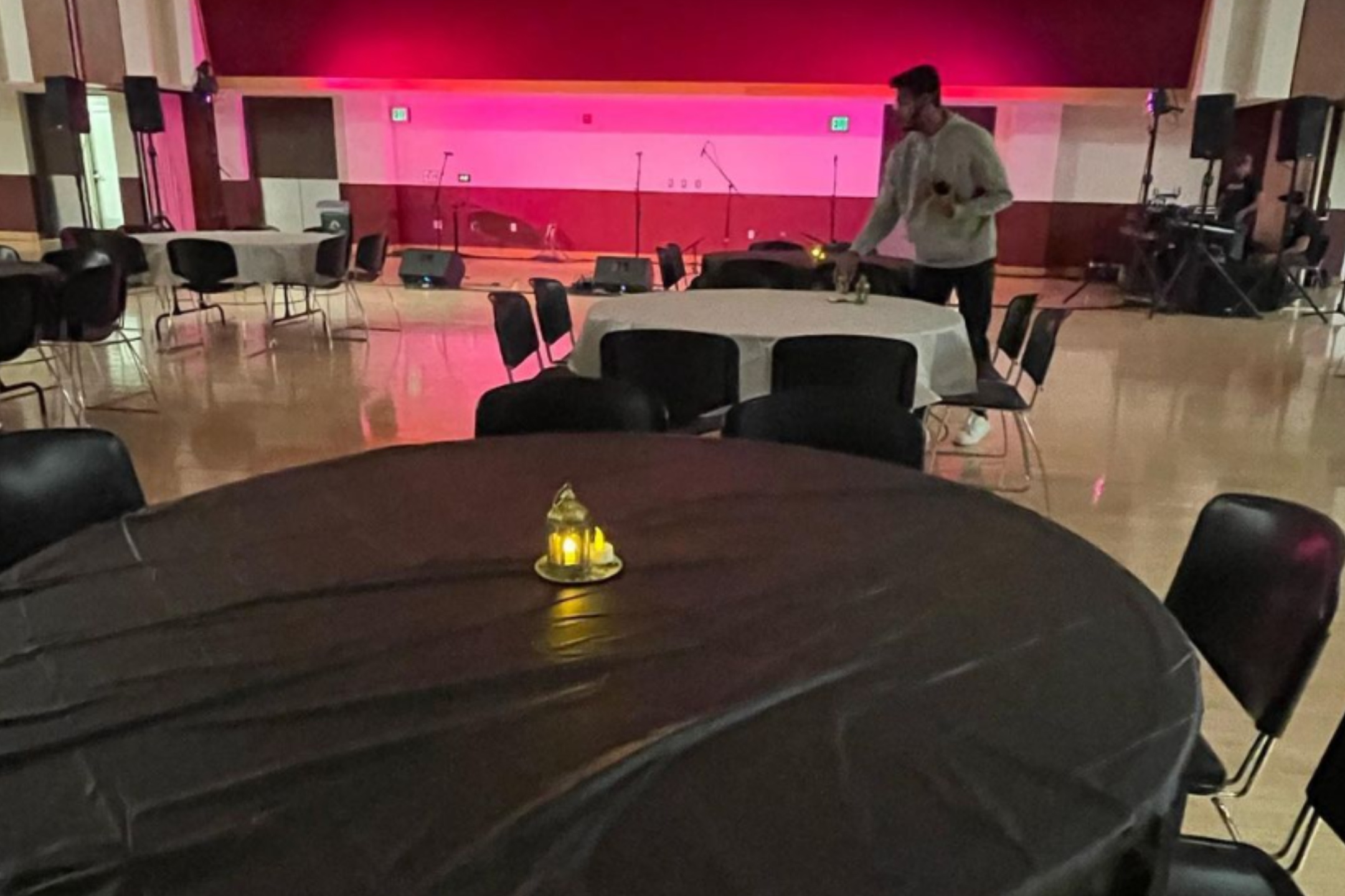 he image depicts a medium-sized event hall with polished, light-colored floors and round tables set up for an event. The room is dimly lit, and the stage at the back is illuminated with a red light, casting a warm glow across the area. Several round tables are covered with black tablecloths, each adorned with a small, lantern-like candleholder holding a glowing candle inside. Chairs are arranged around the tables, predominantly black in color. In the foreground, one of the tables and its candleholder are clearly visible. In the middle ground, a person in a gray hoodie is adjusting items on a table. In the background, the stage has microphones set up, with speakers and various equipment on either side.