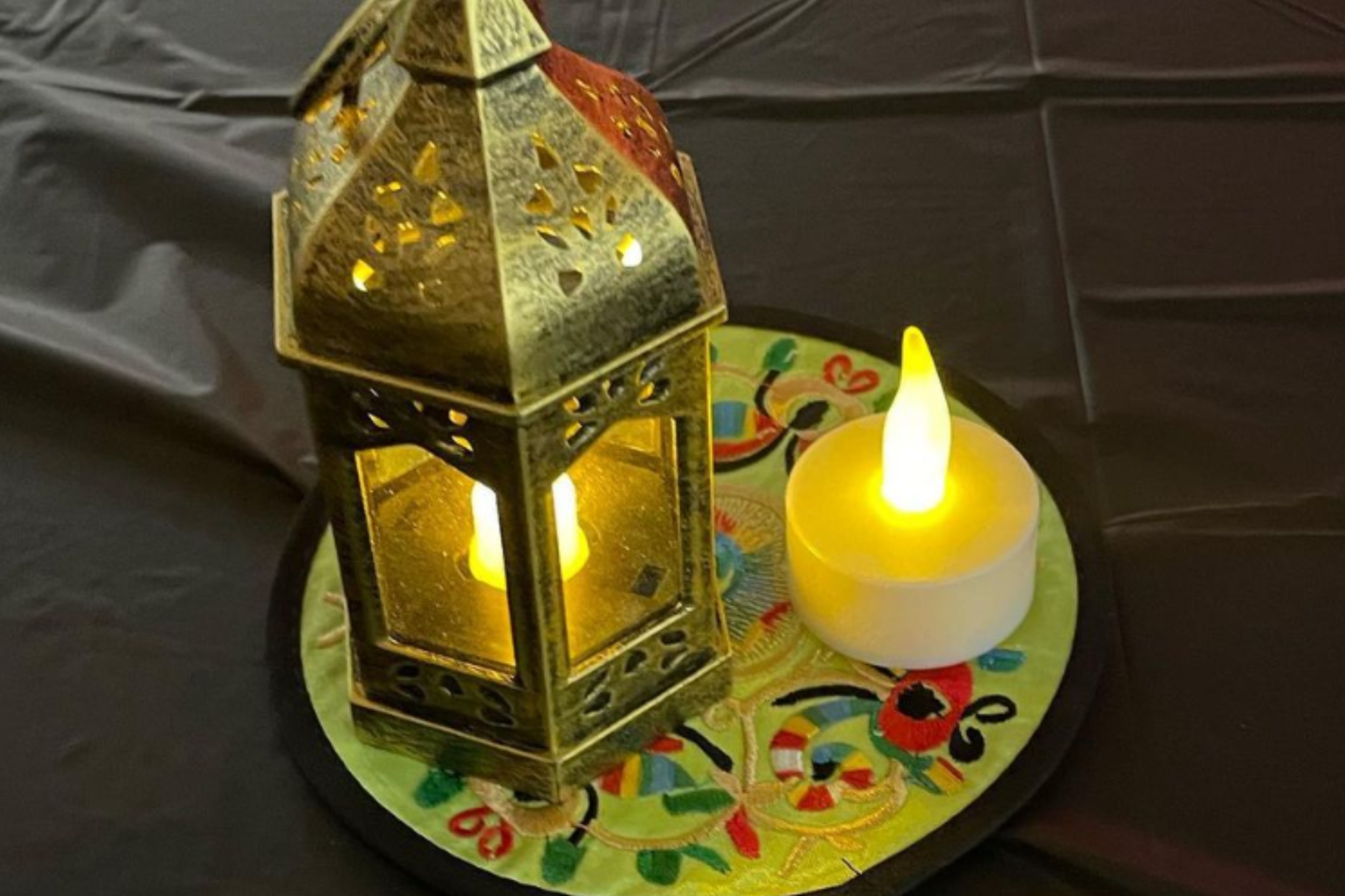 The image showcases a decorative metal lantern and an LED candle placed side by side on a round, colorful base. The lantern is primarily yellowish-gold and features intricate cut-out designs, allowing light to shine through. The LED candle next to it emits a warm yellow glow. Both items are set on a round base with a multicolored, festive design which includes green, red, and blue floral patterns. The background consists of a dark, folded tablecloth.