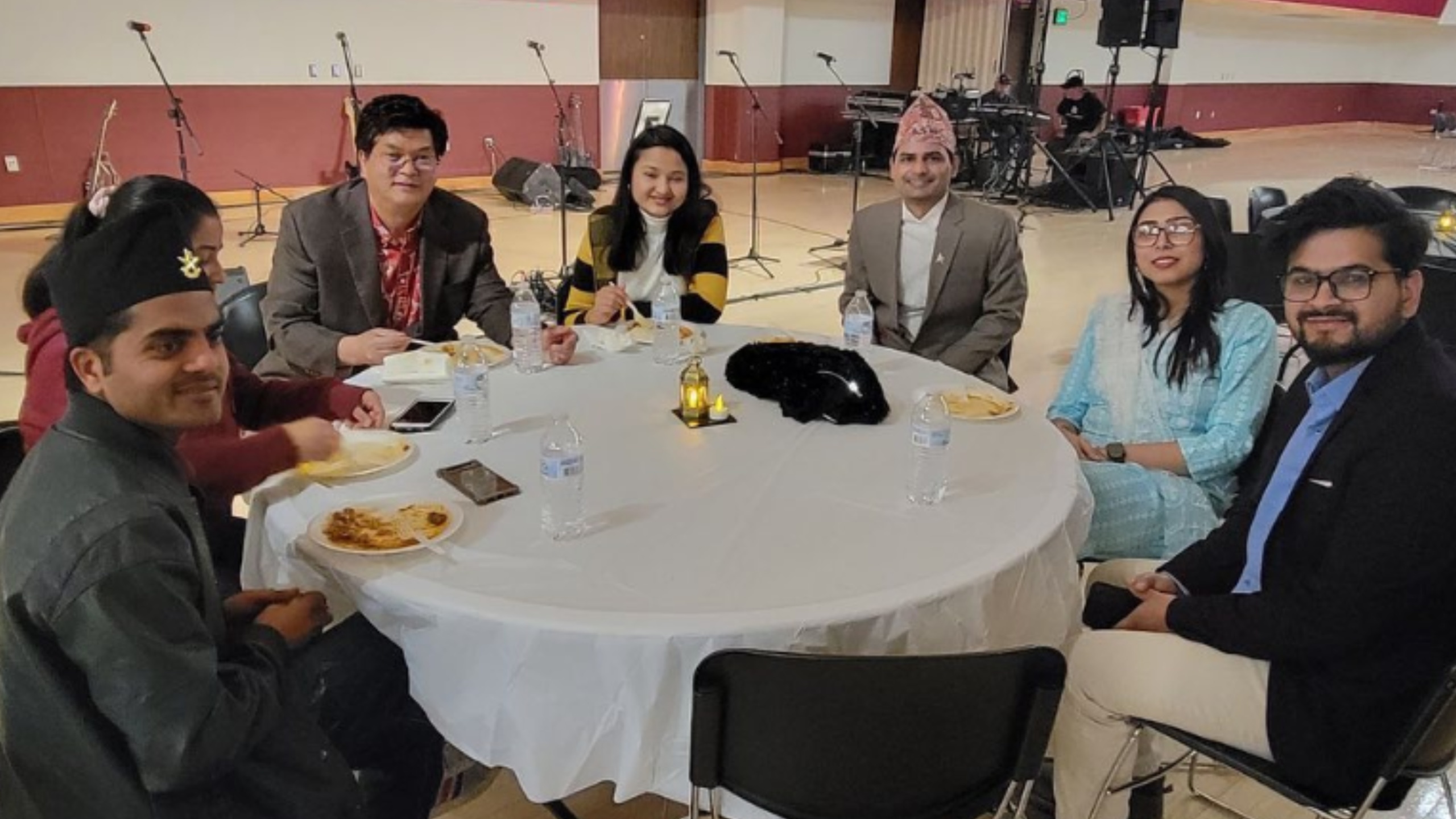 The image shows a group of seven people sitting around a round table at an indoor event. The table is covered with a white tablecloth and has water bottles, plates with food, a small decorative item with a candle, and a black cloth on it. The group appears to be enjoying a meal. The background features a stage setup with musical instruments, microphones, and other equipment. The venue has a polished floor and a mix of red and white walls. The individuals are dressed in various traditional and modern attire and appear relaxed and engaged with one another.