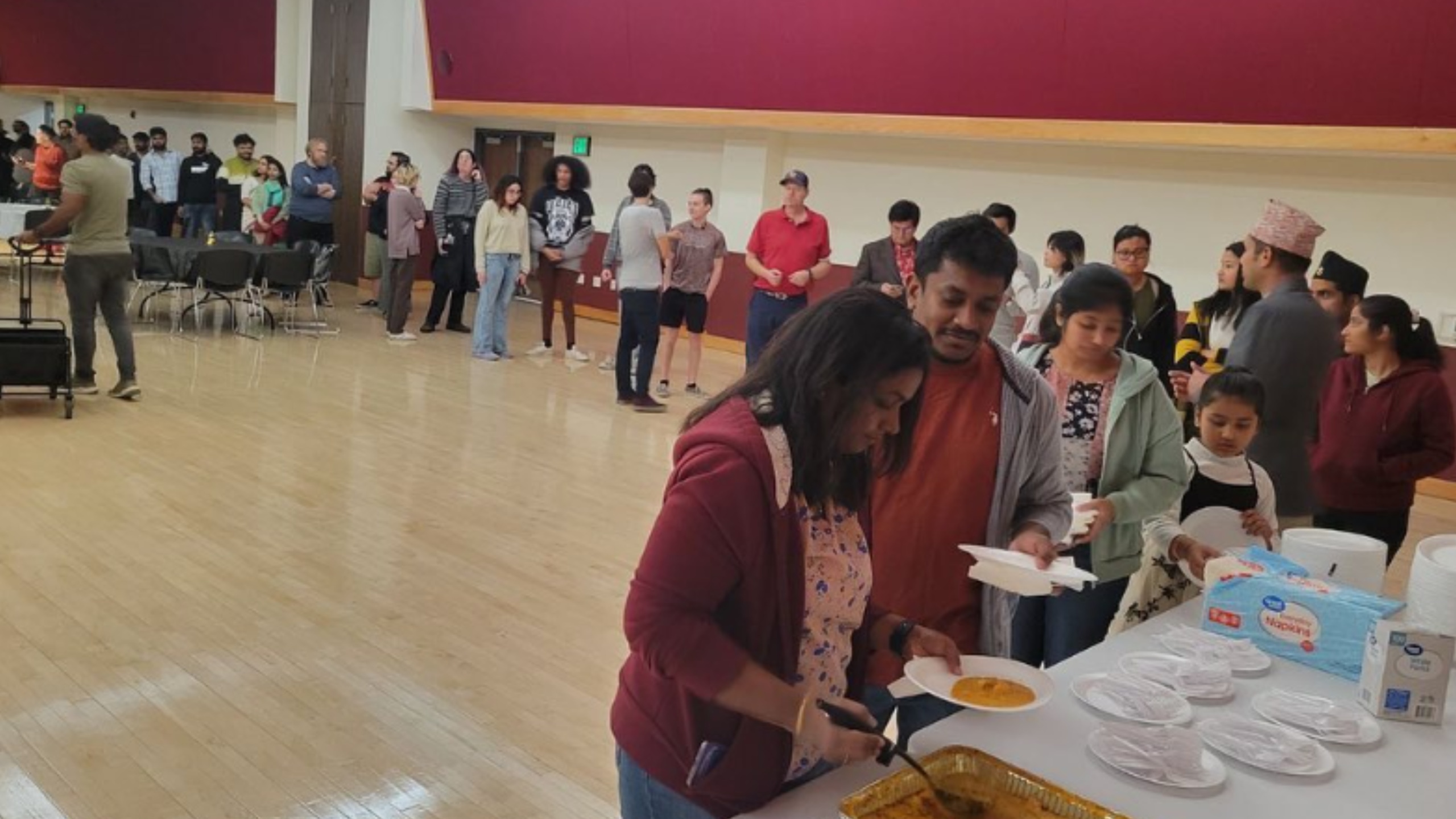 The image depicts a group of people gathered indoors, seemingly at a communal event or gathering taking place in a large hall with a light wooden floor. In the foreground, a woman in a red jacket is serving food from a large aluminum tray onto a white plate. Standing next to her is a man holding a white plate, followed by other adults and children in a queue, waiting to be served. To the right, there is a table with stacks of disposable plates, napkins, and cups. In the background, more individuals can be seen mingling and some others are seated around round tables and chairs placed along the length of the room. The walls are painted in a combination of red and beige and the overall atmosphere appears casual and relaxed.