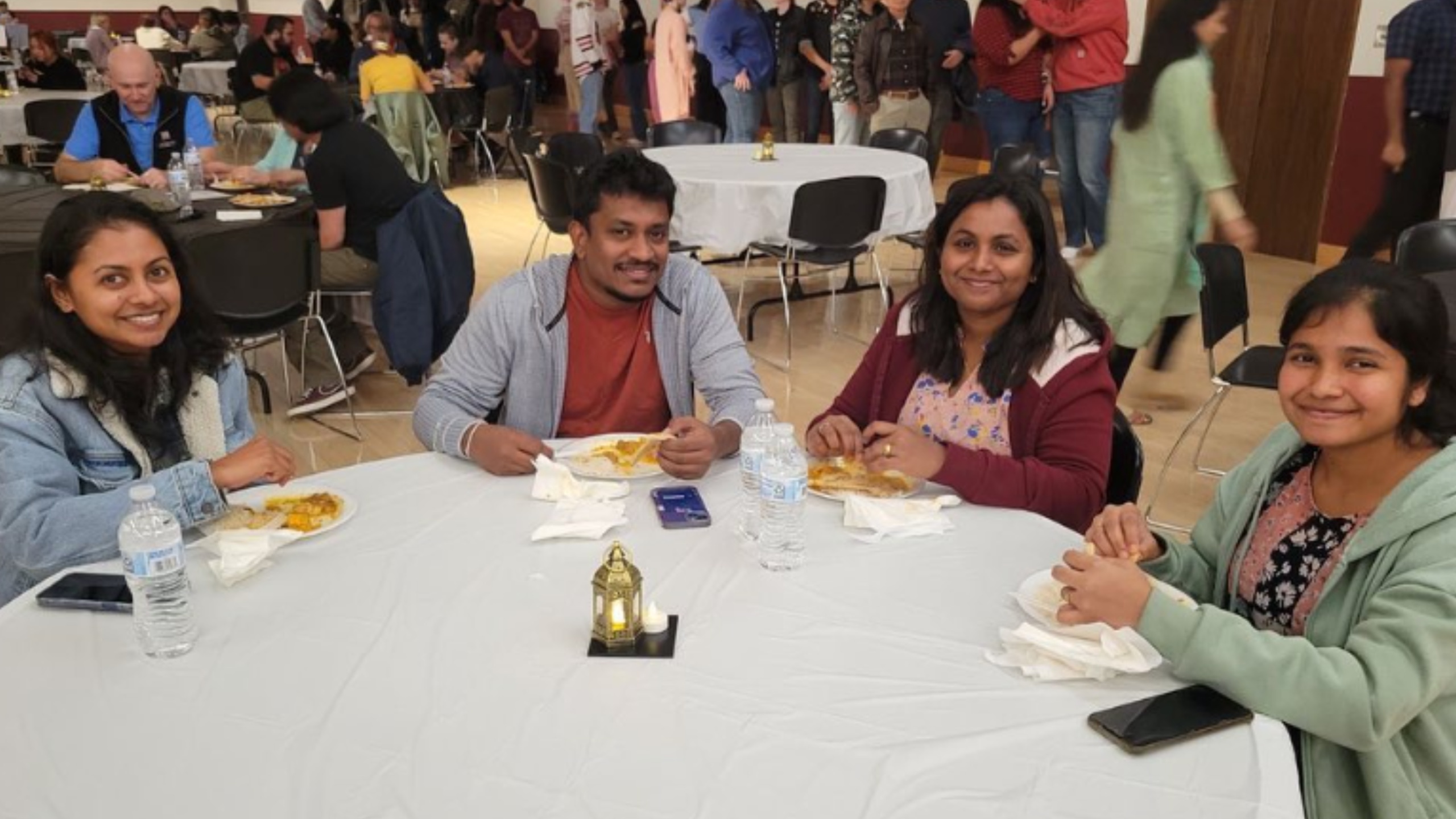 The image shows a group of four people seated at a round table covered with a white tablecloth. The table features four plastic water bottles, some paper napkins, and a small decorative lantern with a lit candle inside. The individuals appear to be enjoying a meal; they have plates of food in front of them. The setting is indoors, possibly in a large hall or cafeteria. In the background, there are multiple other tables with groups of people sitting and engaged in conversations. The room has a lively atmosphere. The floor is wooden, and some people are seen standing, talking, or walking around.