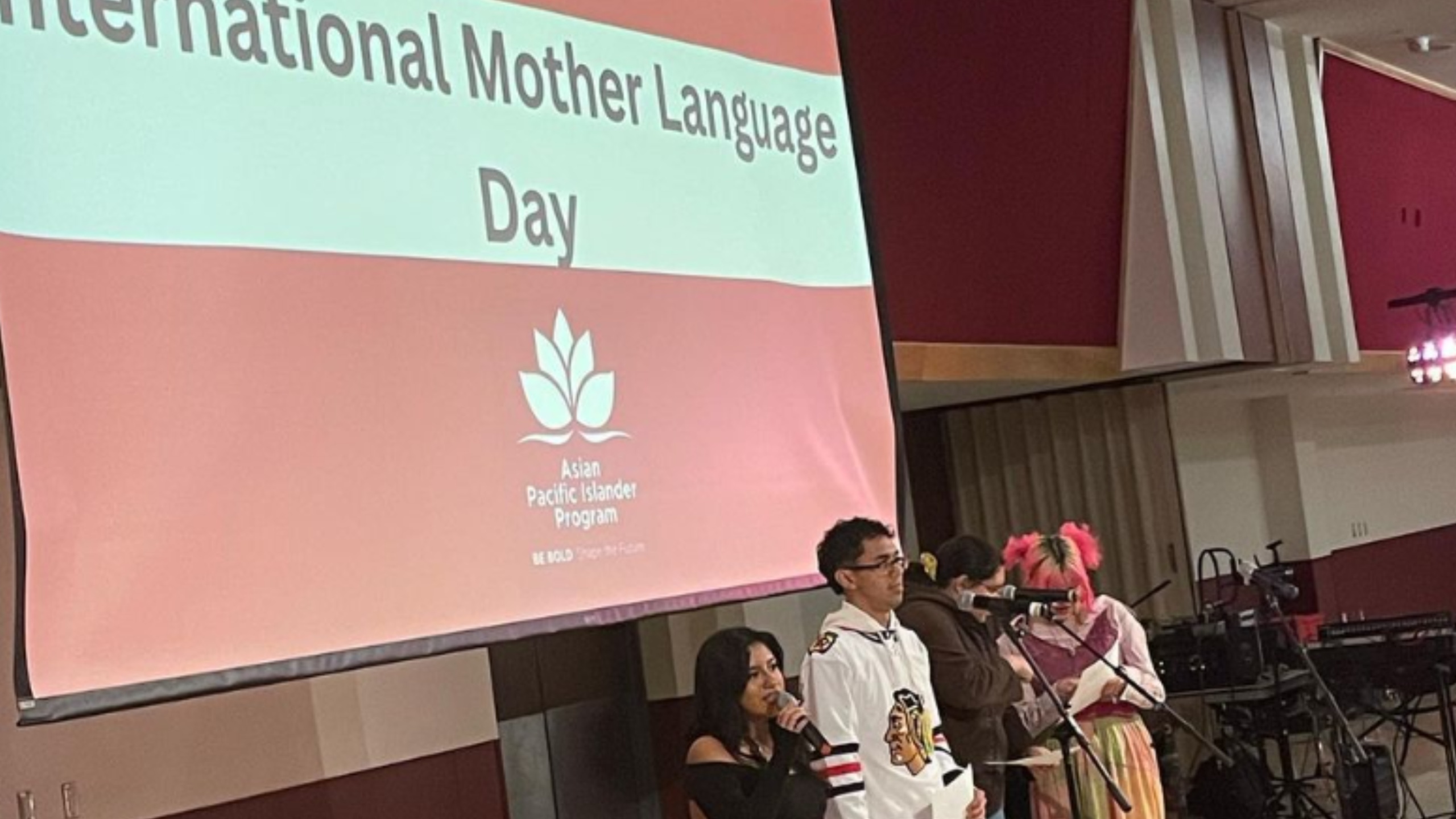 he image portrays a group of people on stage at an event celebrating "International Mother Language Day." A large screen in the background displays the event title in bold letters against a red background, featuring a white emblem of a lotus flower and text below it. Four individuals are on stage, each standing in front of a microphone. The person nearest to the camera is a woman with long dark hair dressed in a black outfit, speaking into a microphone. Beside her is a man wearing glasses and a white sports jersey with a logo. Two more people are seen further in the background, one with pink hair and a colorful outfit, reading from a script, and another wearing a brown jacket and a mask. The setting is a room with maroon walls and stage equipment, including microphones and stands, visible in the background.