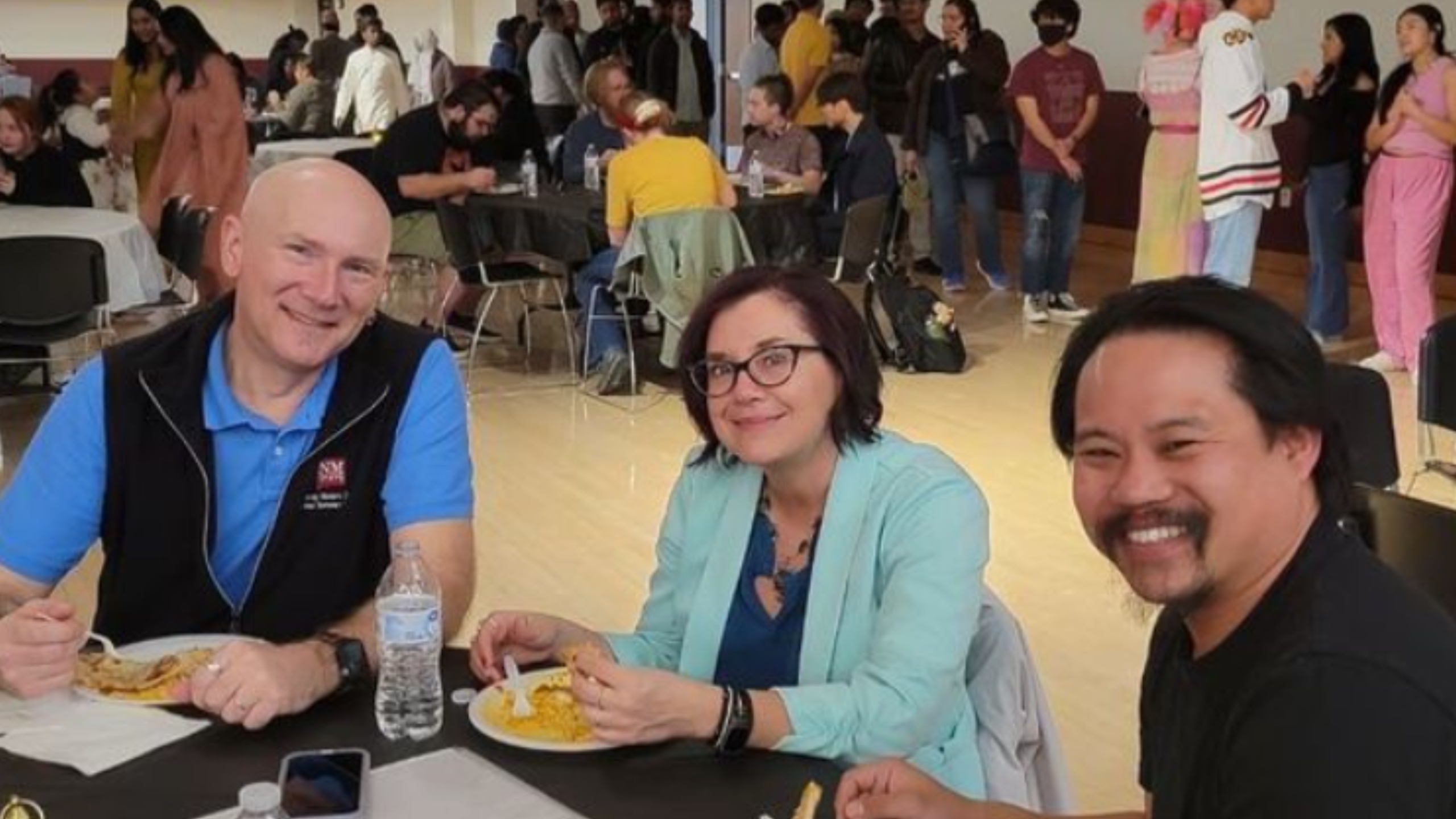  The image shows three people sitting at a table in a crowded indoor event, likely a social gathering or celebration. They are in the foreground of the image, smiling at the camera while eating from their plates. The man on the left is bald, wearing a blue polo shirt and a black vest, holding a forkful of food. In front of him is a water bottle and a smartphone lying on the table. The woman in the middle has short dark hair, is wearing glasses, and is dressed in a light blue blazer over a blue top. She is holding a piece of food with one hand and smiling. The man on the right has black hair and a mustache, wearing a black shirt, also smiling. The background depicts other attendees sitting at tables and standing in groups, with some engaged in conversation. The atmosphere appears lively and social.