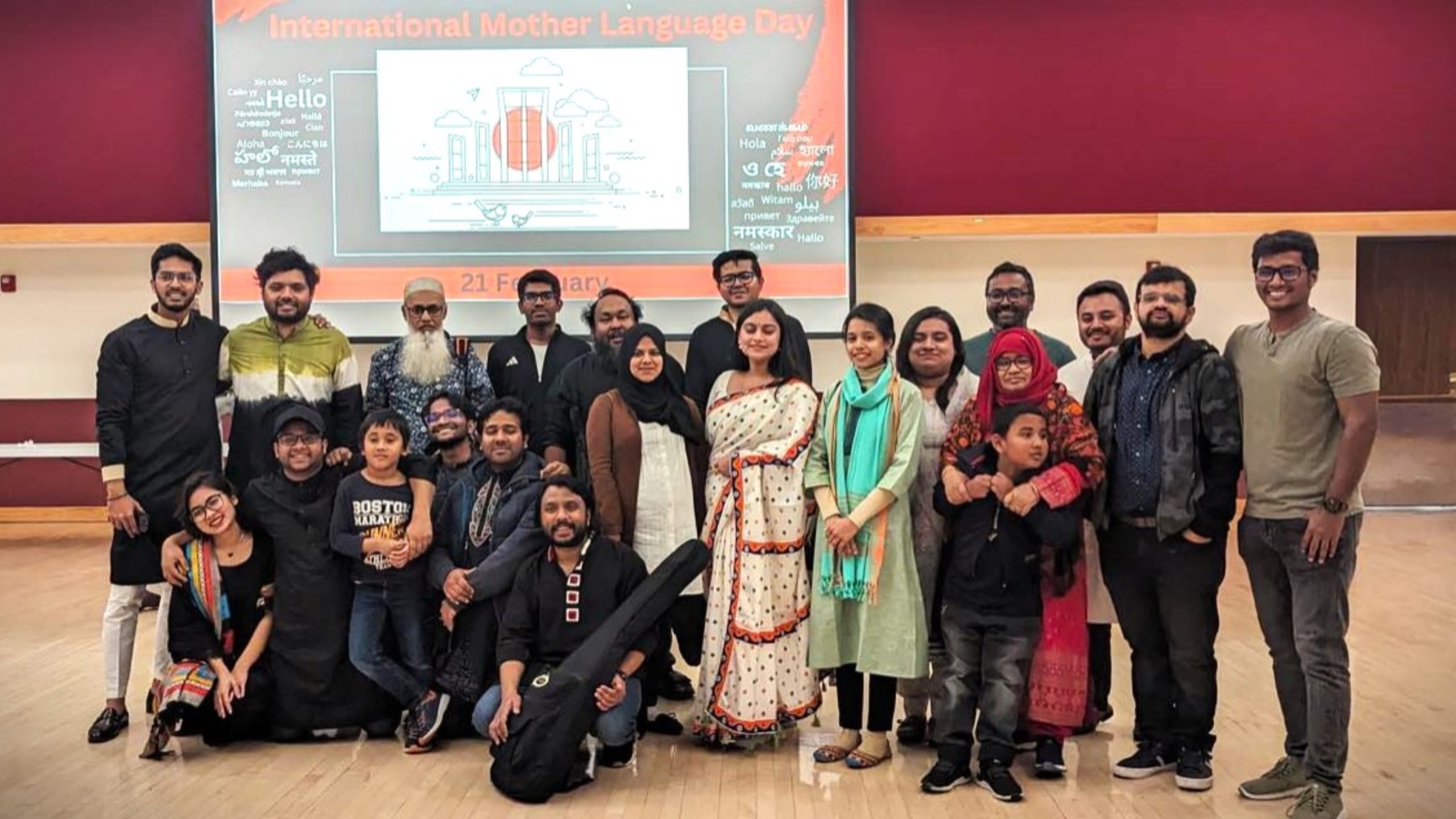 The image depicts a group of twenty-one people standing and sitting together in a large room. The group includes men, women, and children of different ages. The setting appears to be an event or celebration, as indicated by the large screen behind them which displays a colorful graphic and text. The people in the image are mostly wearing casual to semi-formal attire; some individuals are dressed in traditional clothing.