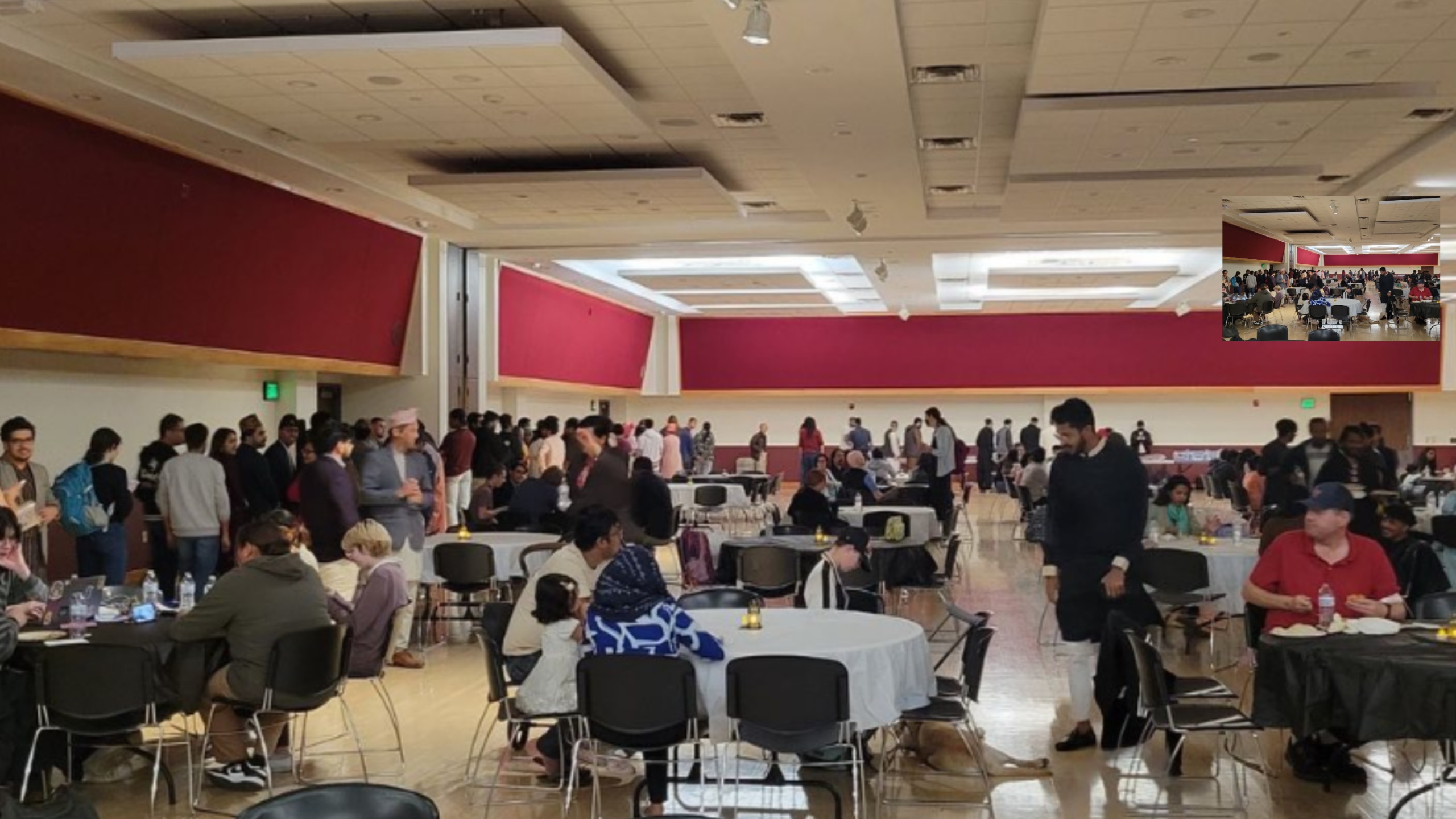 The image depicts an indoor event held in a large, well-lit room with light-colored walls and a polished wooden floor. There are numerous round tables draped with white and black tablecloths, surrounded by black chairs. Many people are present, some seated at the tables engaged in conversation or eating, while others are standing in line along the room's side.  The ceiling features a grid of recessed panels with embedded lighting. The walls have broad, vivid red panels providing a striking contrast to the otherwise neutral-toned interior. The overall atmosphere appears lively and social, indicative of a gathering or social function.