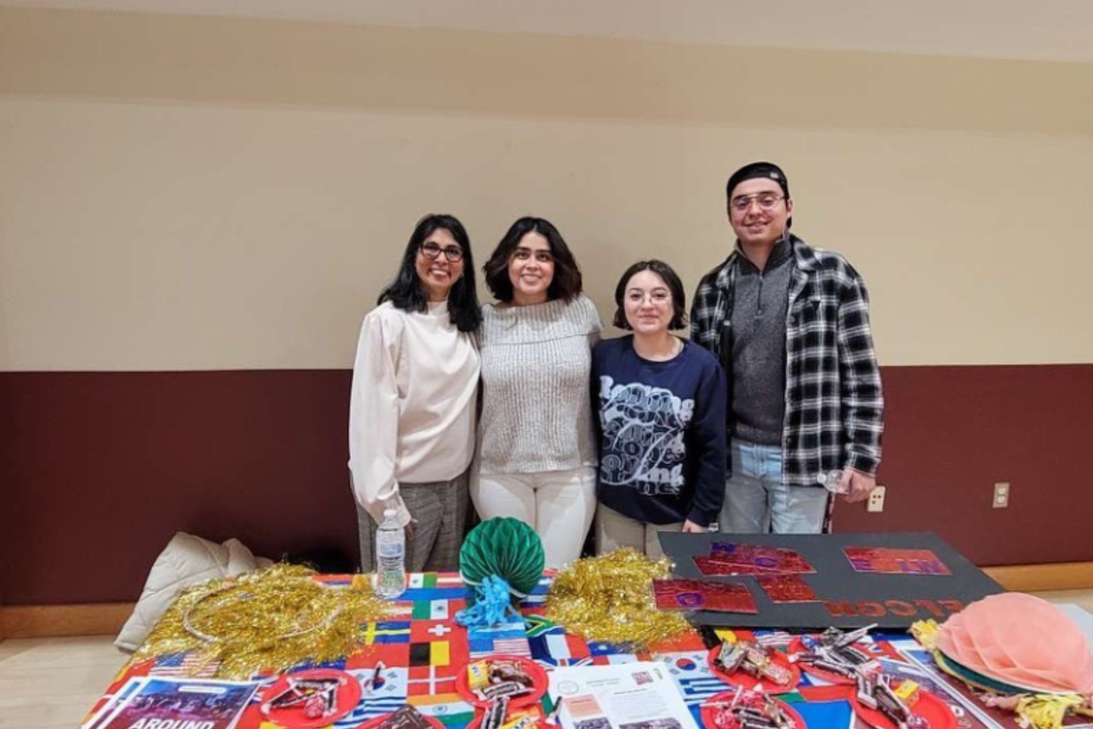 A group of four individuals is standing behind a decorated table. The table is covered with a vibrant tablecloth featuring a pattern of various international flags. On the table, there are several items including a plastic water bottle, gold tinsel, a green paper decoration, pamphlets, candy packets, and a few arts and crafts materials. Behind the table, there is a plain wall with two horizontal color bands, beige on the top and maroon on the bottom.  The first person on the left is wearing glasses and a white blouse. The second person in the center-left has shoulder-length dark hair and is wearing a light-colored sweater. The third individual in the center-right has short dark hair and is wearing a navy blue sweatshirt with white textual graphics. The fourth and last individual is wearing glasses, a black and white checkered jacket over a gray shirt, light blue jeans, and a black cap worn backward.