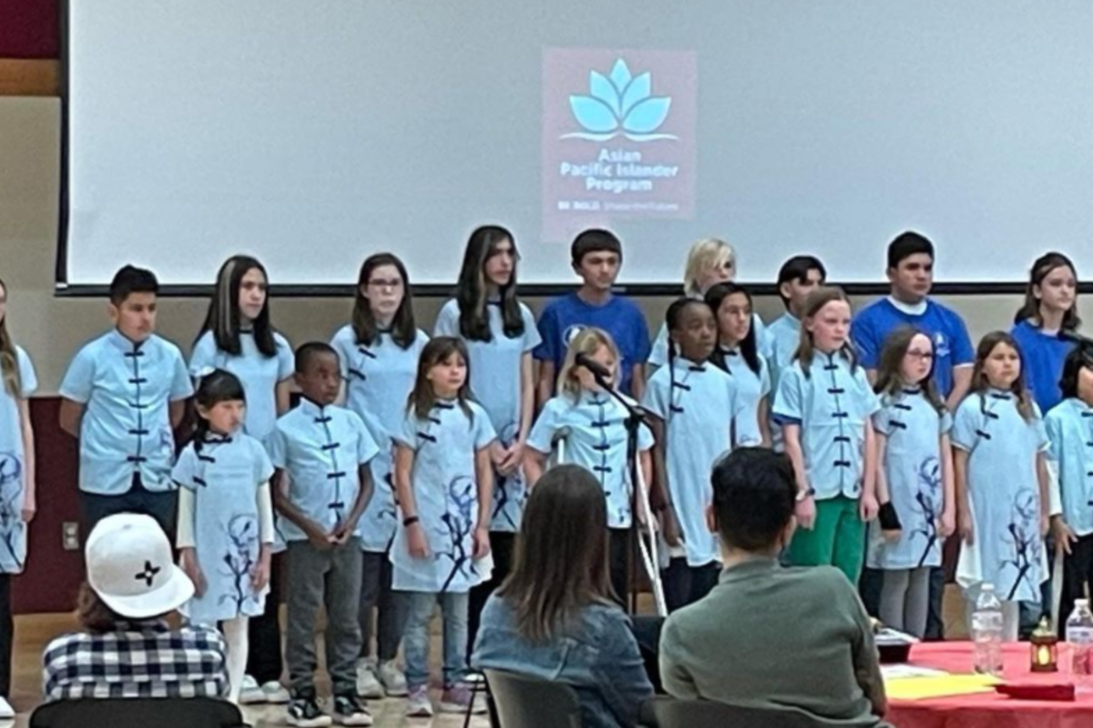  The image depicts a group of children standing on a stage in a school or community center. They are arranged in rows and appear to be ready to perform or have just performed. Most of the children are wearing light blue traditional dresses with dark blue trim and a floral design. In front of the group, there is a microphone stand. A screen behind the children shows a logo with a lotus flower and text that reads "Asian Pacific Islander Program." There is an audience seated in front of the stage, including a person in a white cap and another in a denim jacket. Various items are placed on tables in the foreground, including a water bottle and a few decorations.