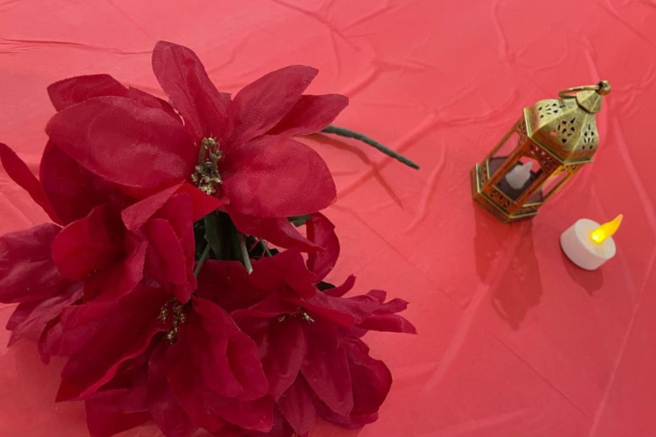 The image shows a decorative setup on a red tablecloth. Positioned in the center-left are vibrant, large red fabric flowers with green stems. To the right of the flowers, there is a small, golden metal lantern with intricate cut-out designs and a glass panel. Beside the lantern is a small, battery-operated tea light candle with a yellow flame-shaped bulb. The tablecloth is slightly wrinkled and matches the red color scheme of the flowers.