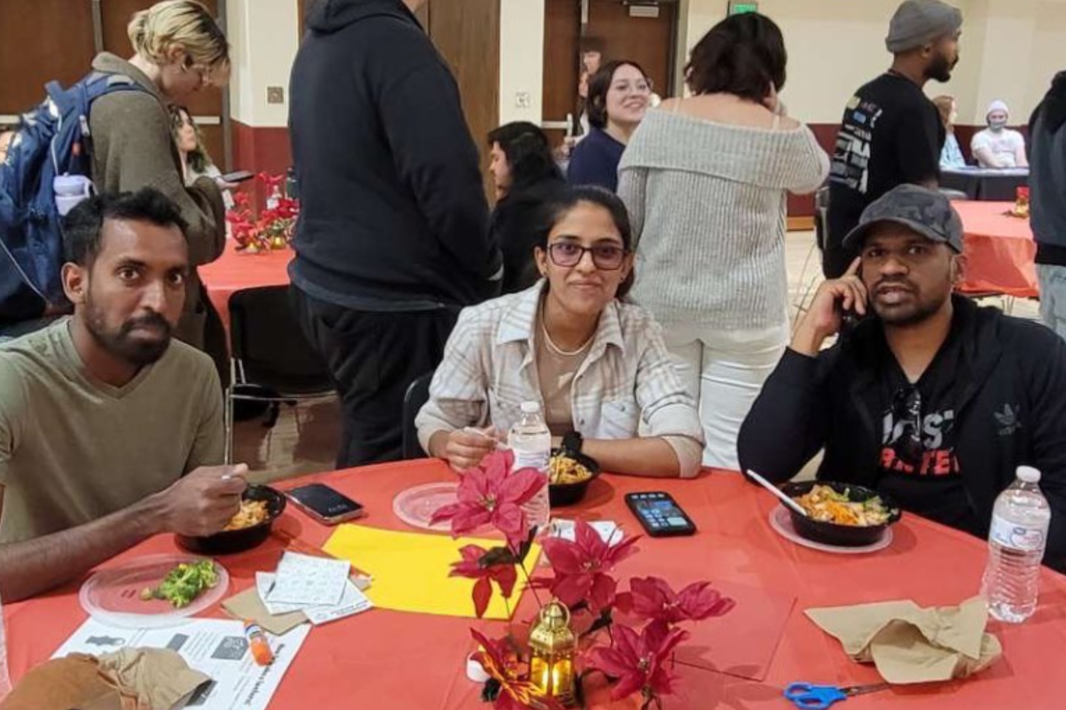 he image captures a group of people in what appears to be a social gathering or event. Three individuals are seated at a round table draped with a red tablecloth, which features a flower arrangement with red blossoms and a small decorative lantern in the center. The person on the left is wearing a grey-green long-sleeve shirt and is holding a fork, eating from a black bowl. The individual in the middle is wearing glasses, a light checkered shirt, and is also eating from a black bowl, with a water bottle on the table. On the right, a person in a black hoodie and a dark cap is holding a phone to their ear, with another black bowl in front of them. Several items like a phone, a plastic container with green vegetables, papers, and a glue stick are scattered around the table. In the background, other people are standing and interacting, and some are seated at other tables.