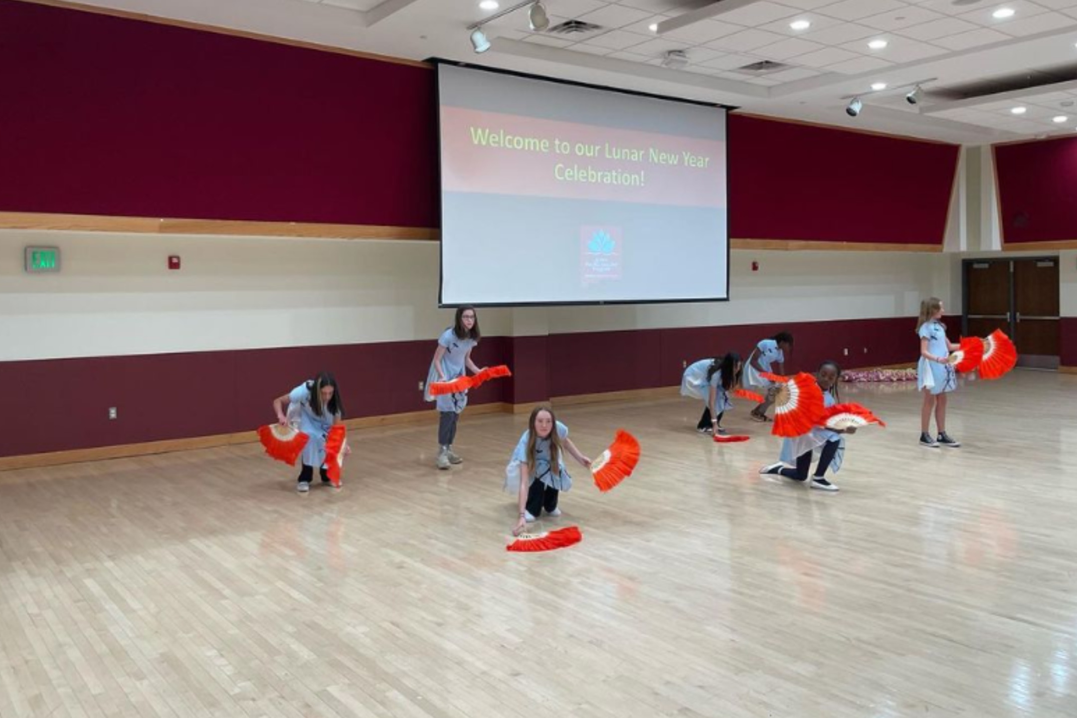 The image depicts a group of seven performers, predominantly composed of young girls, participating in a dance routine to celebrate the Lunar New Year. The dancers are arranged across a spacious gymnasium or multipurpose room, featuring a polished wooden floor and cream-colored walls with a maroon panel running horizontally across the room. The dancers are dressed in light-blue tops and dark pants or skirts. Each of them is holding vibrant orange and white fans, which are prominently displayed as part of the routine.  Above the dancers, a large projection screen displays the text, "Welcome to our Lunar New Year Celebration!" in yellow font. The screen and some ceiling lights are part of the room's modern infrastructure, which also includes recessed lights and sound equipment setups.