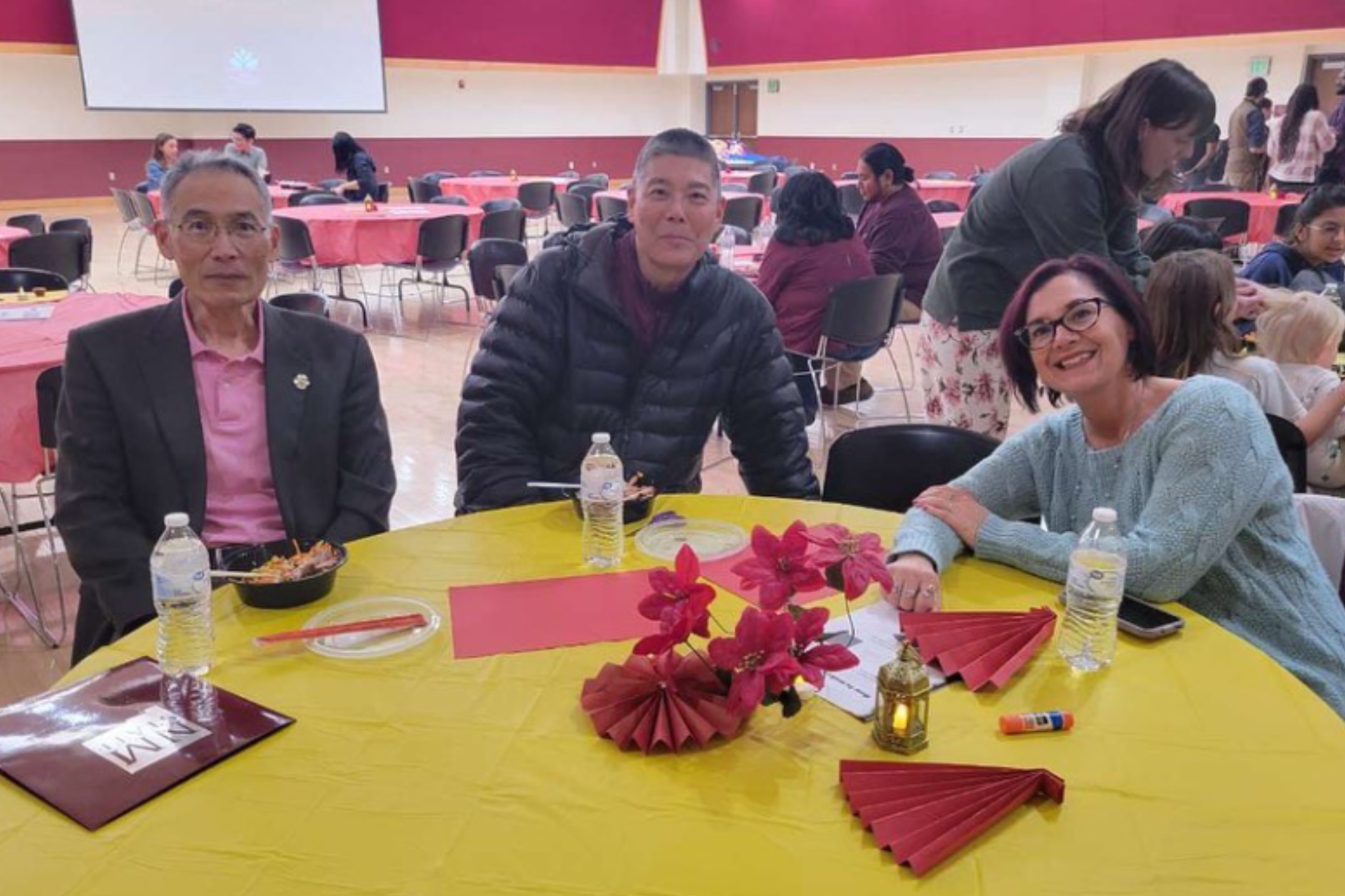 This image depicts three people seated at a round table covered with a yellow tablecloth, participating in an indoor event. The individual on the left is an older man wearing glasses, a dark blazer, and a pink shirt. The person in the center is a middle-aged man wearing a black puffer jacket. On the right is a woman with short hair, wearing glasses and a light blue sweater. The table features two bottles of water, red paper fans, a small decorative lantern, pink artificial flowers, and miscellaneous items including a tube of glue. Around them, the room is filled with similar round tables covered in red or yellow tablecloths, with people engaged in conversations and activities. In the background, there is a large projection screen on a maroon and beige wall.