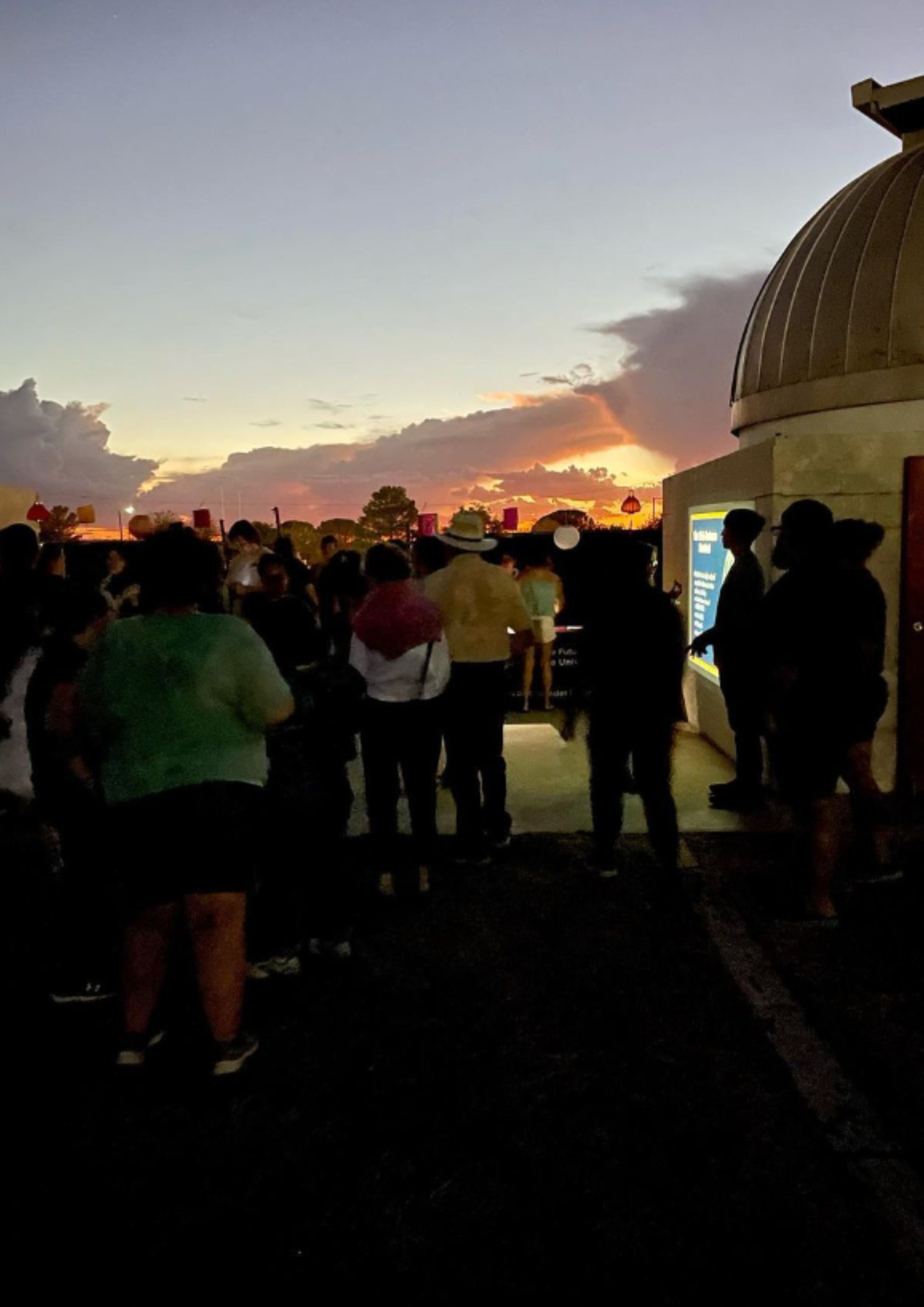 The image depicts a group of people gathered outside at dusk near an observatory dome. The sky is beautifully illuminated with hues of orange, pink, and purple from the setting sun, casting a serene glow over the scene. The observatory dome, located on the right side of the image, is partially in shadow but the structure is clearly identifiable with its rounded metallic roof. To the left, silhouettes of individuals are visible, standing or moving towards the observatory entrance. A bright display panel mounted near the entrance of the observatory emits a blue light, providing some illumination to the nearby individuals. The horizon is dotted with trees and the last glimmers of daylight, creating a picturesque twilight setting.