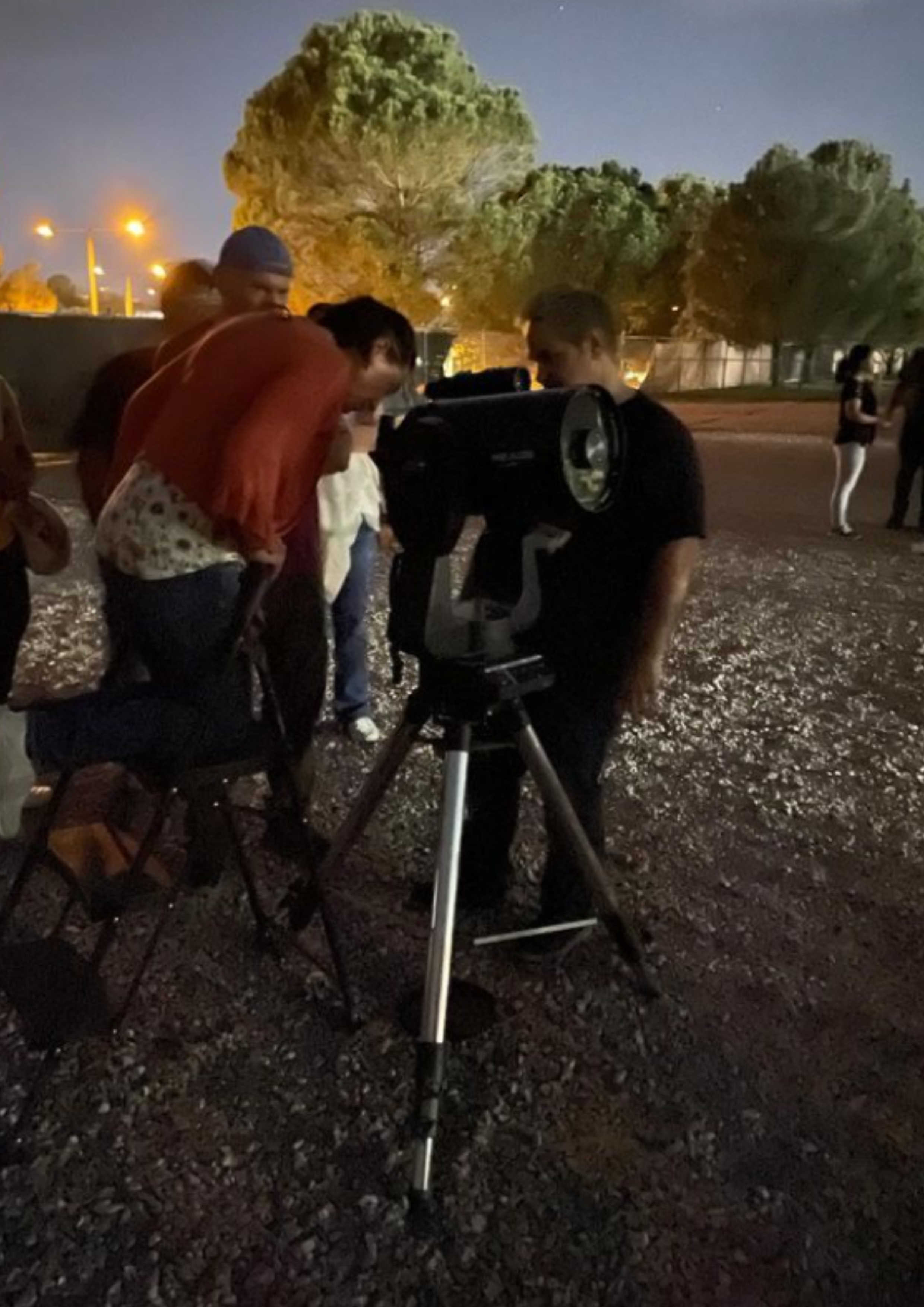 The image portrays a nighttime outdoor scene where several people are gathered around a telescope. The telescope is mounted on a tripod and is positioned on rocky ground. A person in a red top and floral shirt leans on a black chair with one knee to peer into the telescope's eyepiece. Beside them, another individual, dressed in dark clothing, stands next to the telescope, possibly assisting or explaining something. In the background, there are a few other figures standing or conversing. The area is dimly lit with artificial lights illuminating parts of the scene. Large trees with dense foliage are visible behind the people, and the sky is clear with faint stars visible.