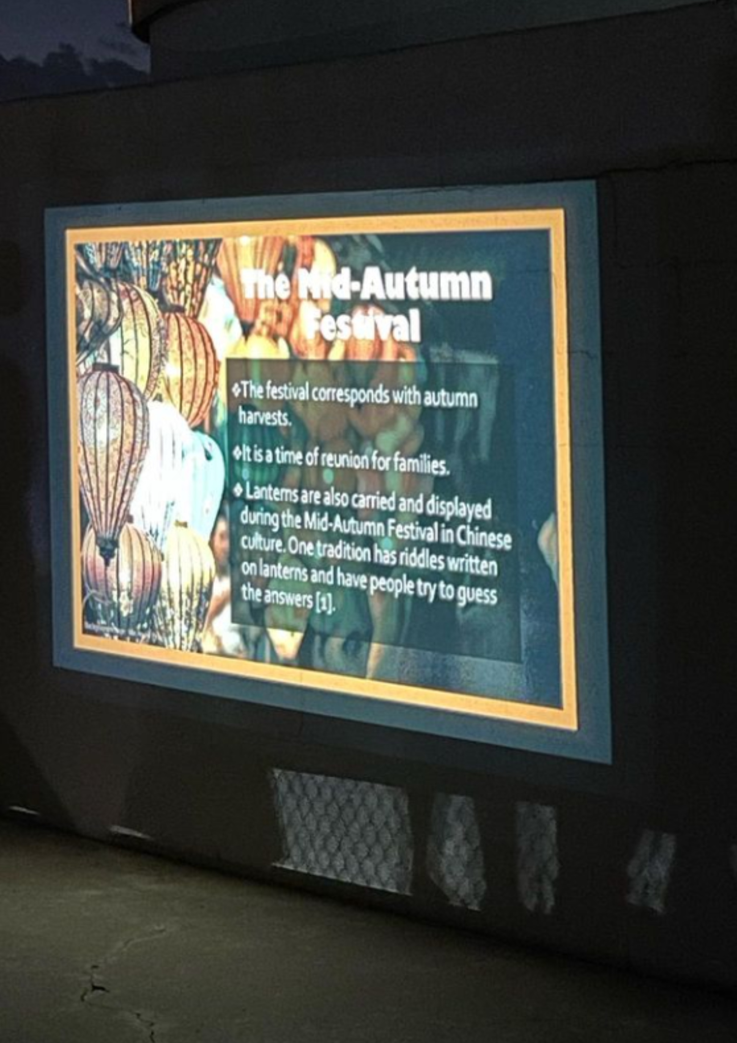 The image is a photograph of a presentation slide projected onto a dark wall. The slide features a title and three textual bullet points on the right side, with an image of several traditionally decorated lanterns on the left. The text is white against a dark background, making it highly legible. The slide border is a thin yellow line. The projected image casts a slight shadow at the bottom, likely due to a mesh pattern below the projector.