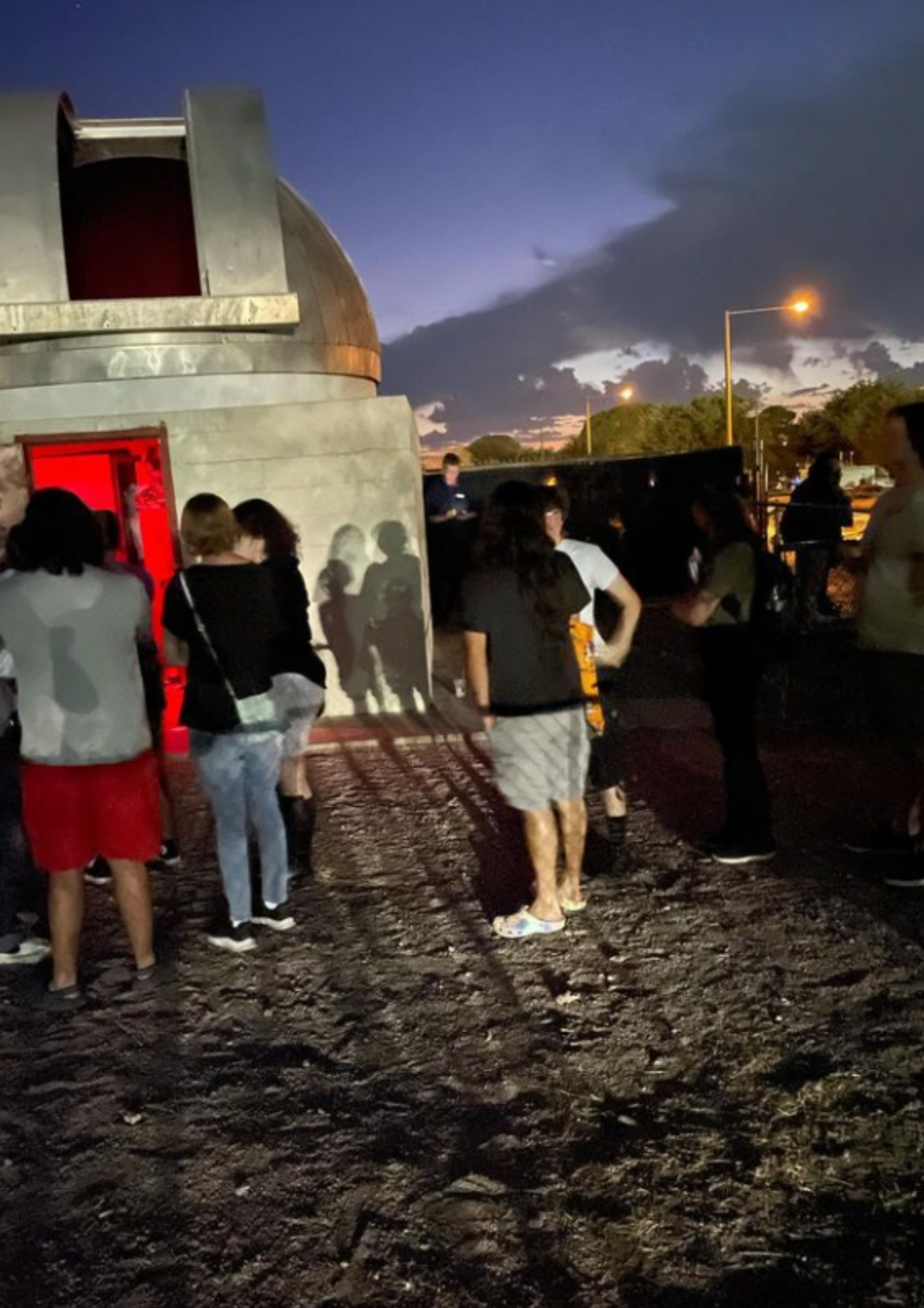 The image shows a group of people gathered in front of a small observatory building at dusk. The observatory features a gray dome with a partially open slit revealing a red-lit interior. The darkening sky has clouds visible, with remnants of a sunset with some orange tints near the horizon and a few streetlights beginning to illuminate. The ground is covered with dirt and small rocks. The individuals in the group are standing in informal clusters, some facing the door of the observatory while others are turned towards each other, engaged in conversation. Shadows of the people are cast on the observatory building from the light above the entrance.