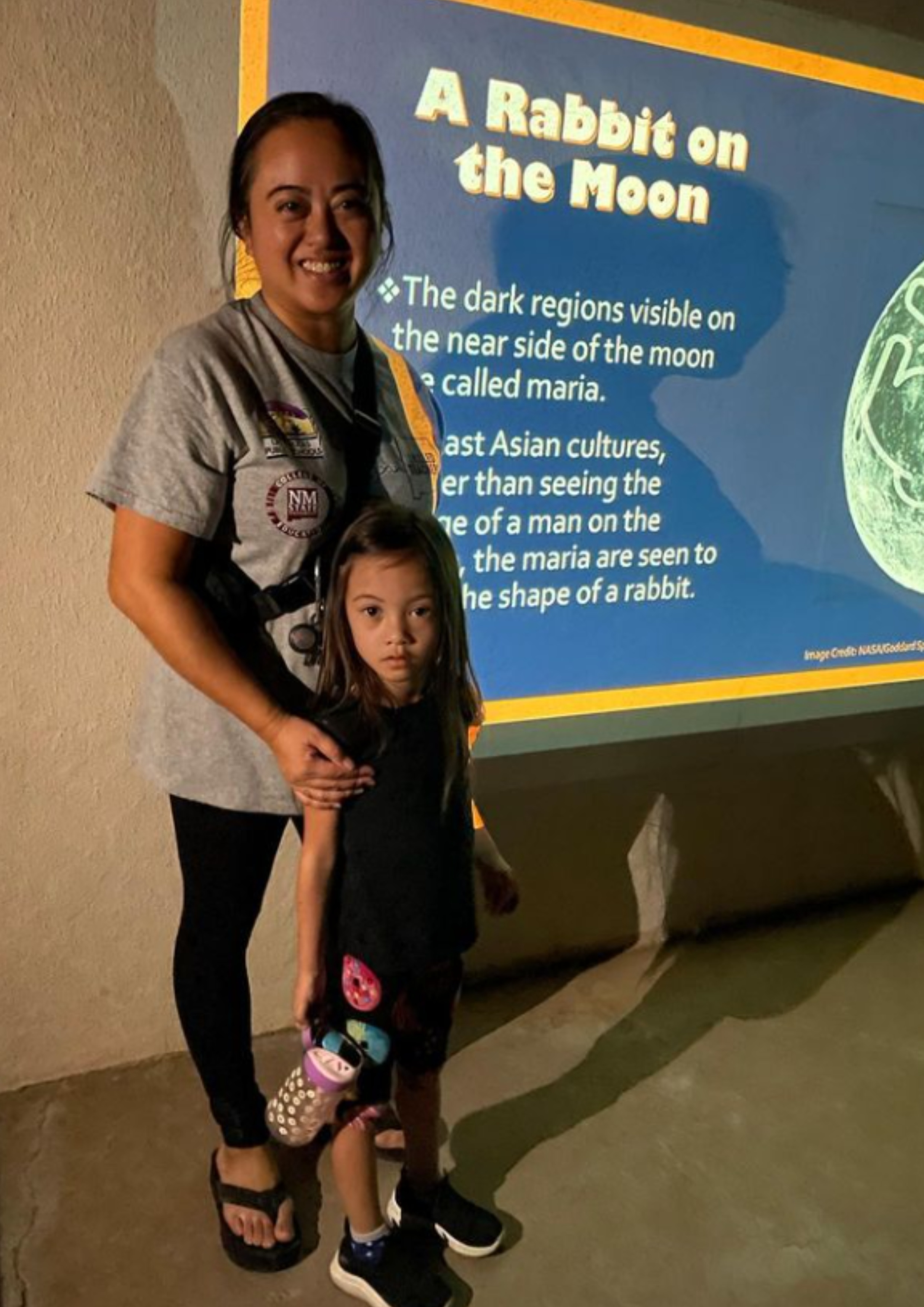 The image captures an adult and a child standing indoors in front of a projected screen displaying information about lunar features. The adult, on the left, is wearing a light-gray T-shirt with badges and black pants, holding the child with their right arm. The child, on the right, is looking directly at the camera, dressed in a black T-shirt and colorful shorts, and holds a polka-dot water bottle in their right hand. The floor appears to be concrete, and the wall behind them is plain taupe.  The projected screen to their right has a dark blue background with a title and text in yellow and white fonts. The title reads "A Rabbit on the Moon." An image of a lunar surface can be seen as part of the projection on the right side.