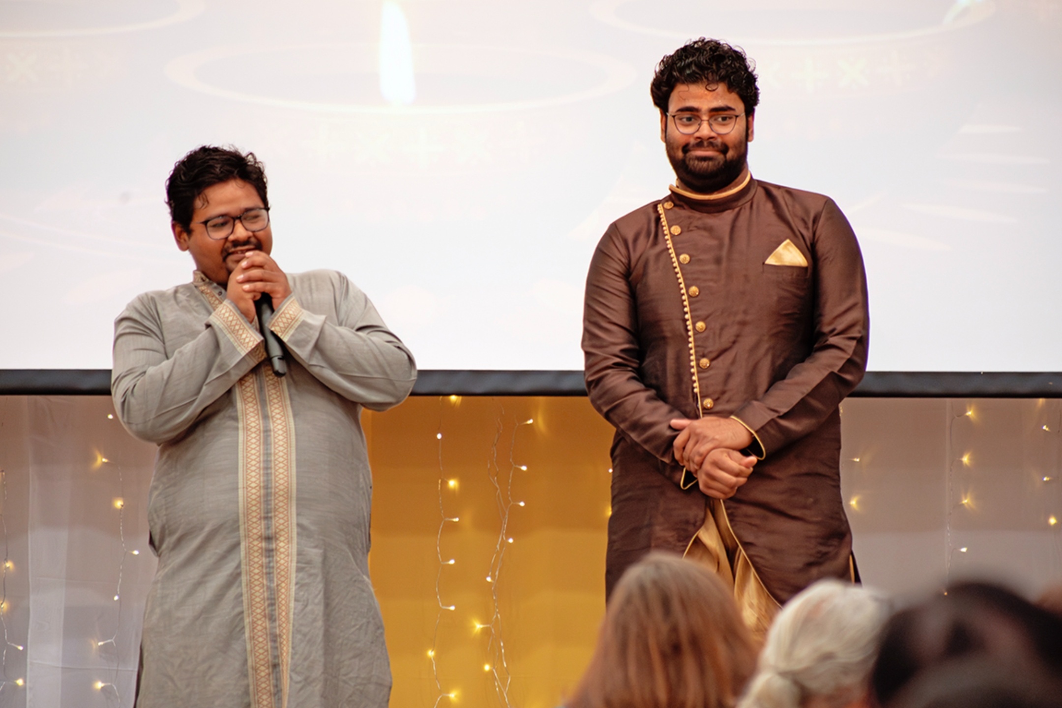 The image shows two men standing side by side on a stage or raised platform. Both men are wearing traditional South Asian attire. The man on the left is holding a microphone with both hands, appearing to be in the middle of speaking. He is dressed in a light gray kurta with intricate embroidery down the front and on the cuffs. He wears glasses and has short, dark hair. The man on the right is standing with his hands clasped in front of him, looking forward. He is dressed in a dark brown sherwani with gold accents, including a decorative chain and a pocket square. He also wears glasses and has short, dark hair. The background is a light-colored curtain with small fairy lights and an image of a lit oil lamp projected on a screen above and behind them. The heads of a few audience members are visible at the bottom of the image.