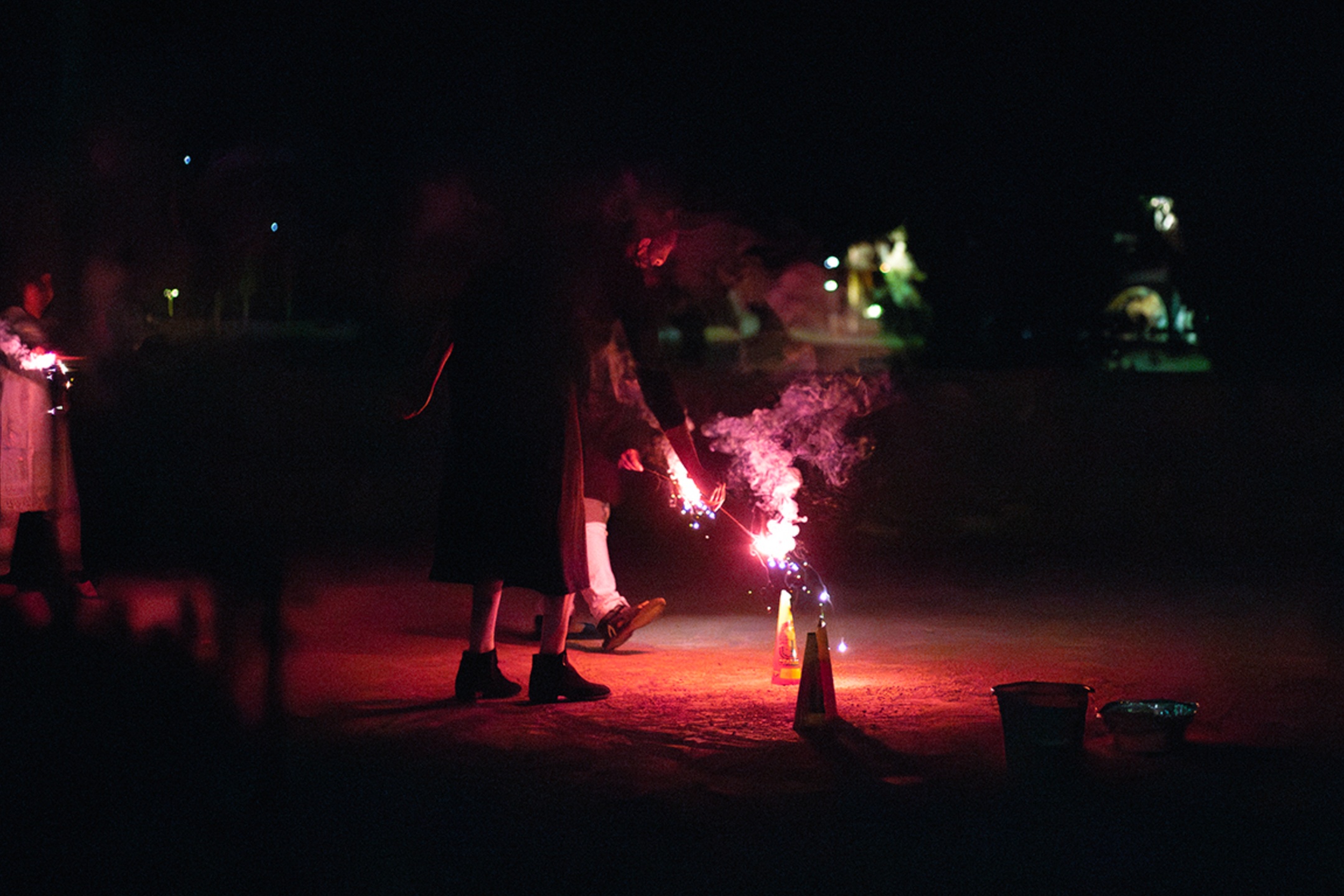 The image shows a night scene featuring individuals handling fireworks. The central focus is on a person dressed in dark clothing, bending over as they light a ground-level firework. The firework emits a bright red and pink light, with sparks and smoke visible. The glow illuminates the surrounding area in a reddish hue. To the right of the person, there are a couple of conical firework casings on the ground. In the background, several other individuals are faintly visible, also holding sparklers. The backdrop is dark with distant street lights and illuminated buildings.