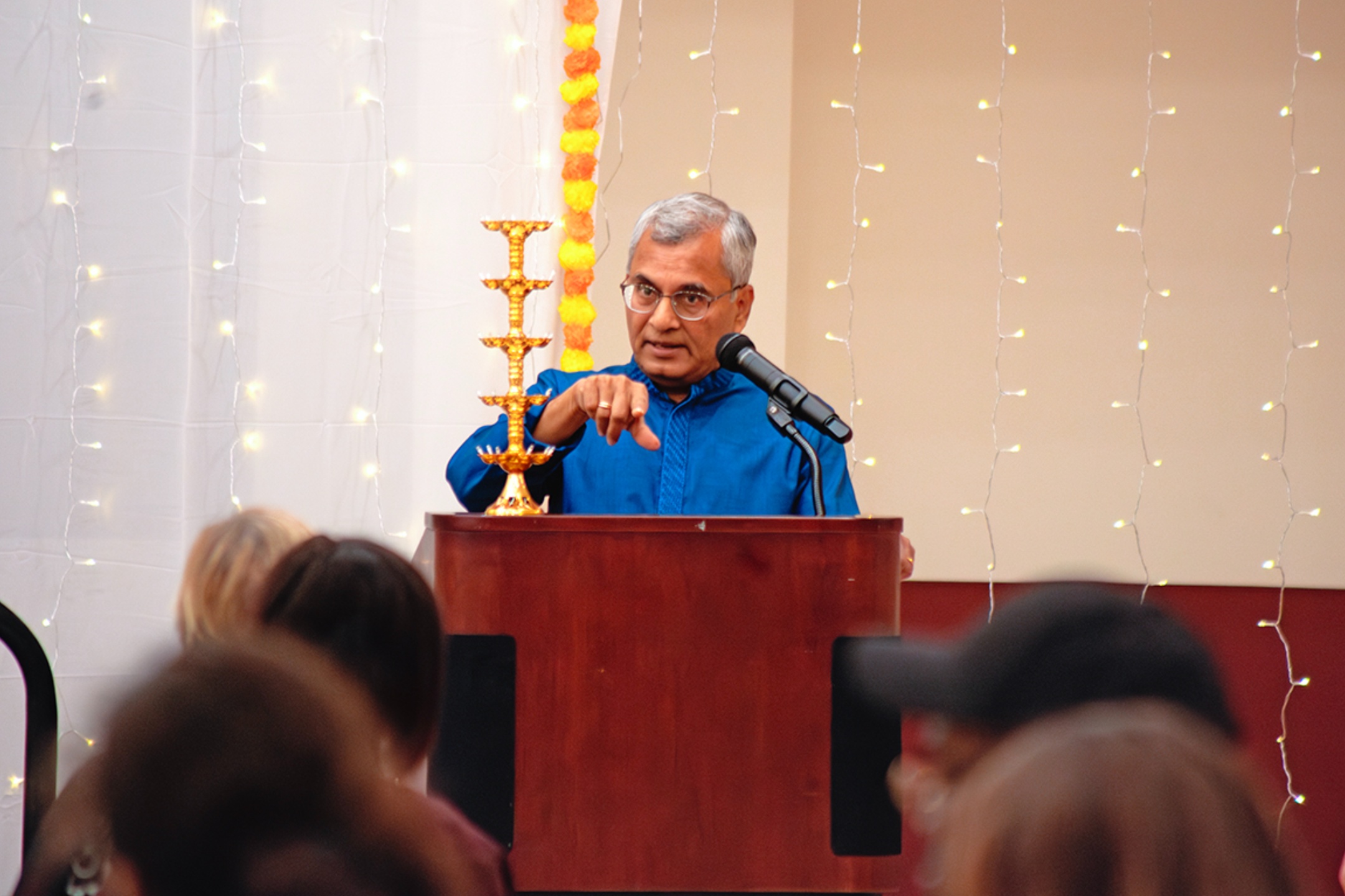 The image shows a man wearing a blue traditional outfit standing behind a wooden podium with a microphone. The man has gray hair, glasses, and is pointing with one finger while speaking. To his left on the podium is a brass multi-tiered oil lamp. In the background, there are white hanging string lights and a vertical garland of orange and yellow flowers creating a festive atmosphere. The audience's heads are partially visible at the bottom of the image.