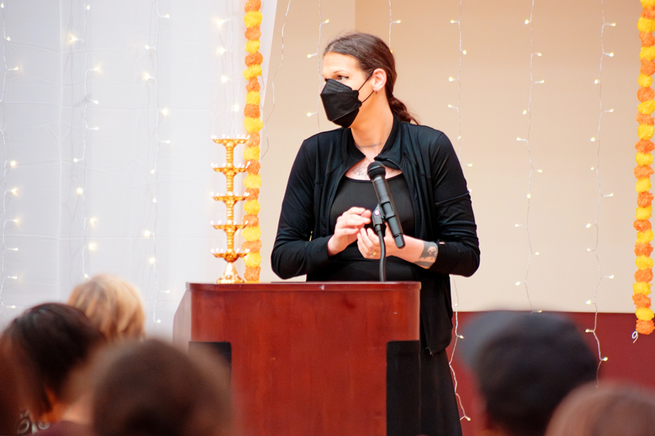 Director of LGBT+ Programs, Zooey Sophia Pook, shows her support for NMSU’s Indian community by attending the Roshni celebration, Sunday, Nov. 5, 2023. The image depicts a person standing behind a wooden podium, speaking into a microphone. They are wearing a black face mask, a black top, and a black jacket. Behind them, the background is adorned with yellow and orange marigold garlands and strings of small white lights. The setting appears to be an indoor event, possibly a formal gathering or a ceremony, inferred by the well-lit and decorated background. The audience is partially visible in the blurred foreground, indicating their attention towards the speaker.