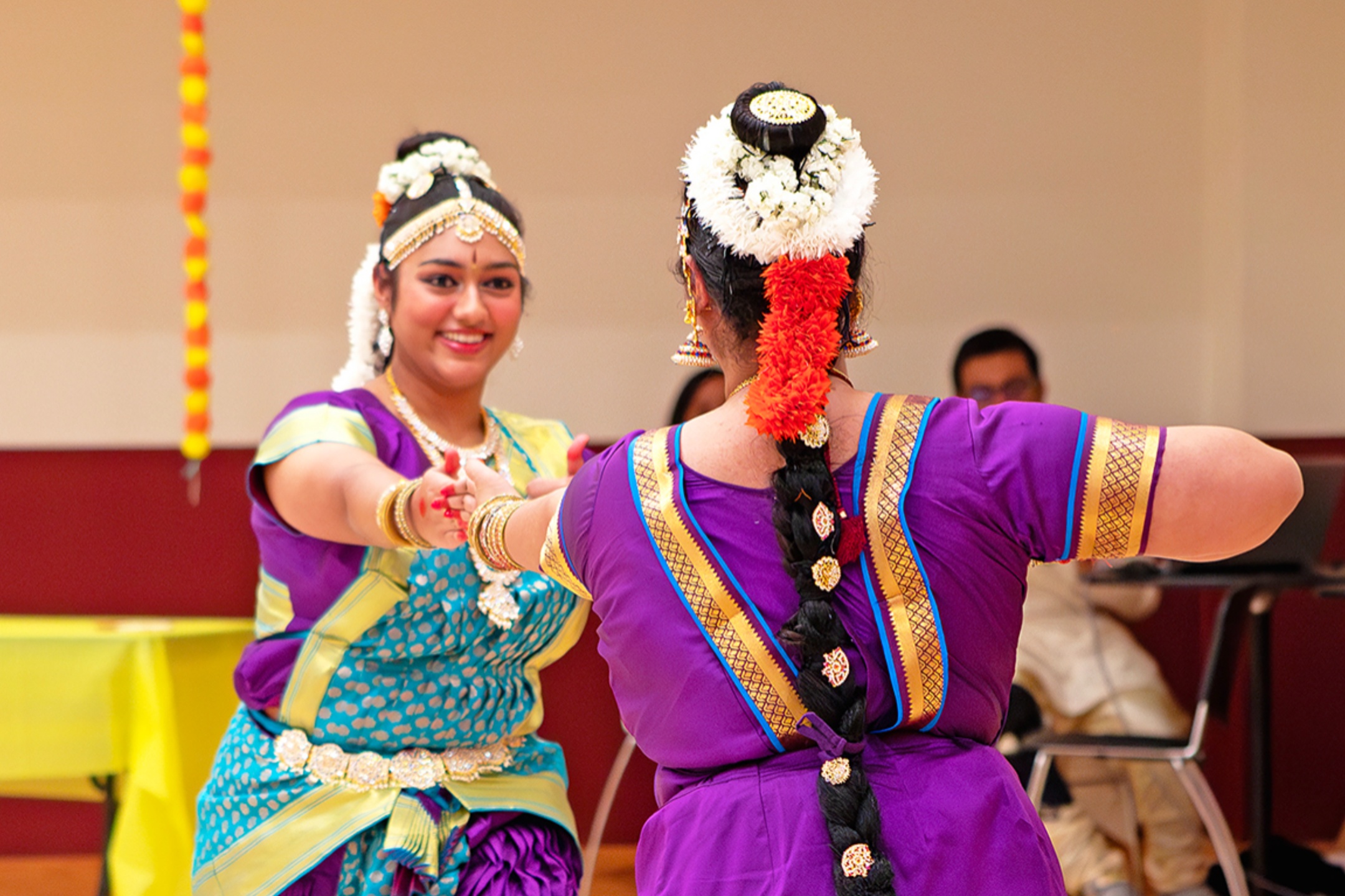 The image captures two individuals engaged in a traditional dance performance. Both are adorned in bright, elaborate attire typical of classical Indian dance. The person facing the camera has a light blue dress with a yellow and green sash, adorned with intricate jewelry including a headpiece, earrings, necklaces, and bangles. The person with their back to the camera wears a vibrant purple and gold outfit with a matching set of jewelry and decorations in her hair, including a floral garland with white and orange flowers. They are holding hands, forming a dance posture. The background is simple, with a red and tan wall and a blurred figure sitting and watching the performance.