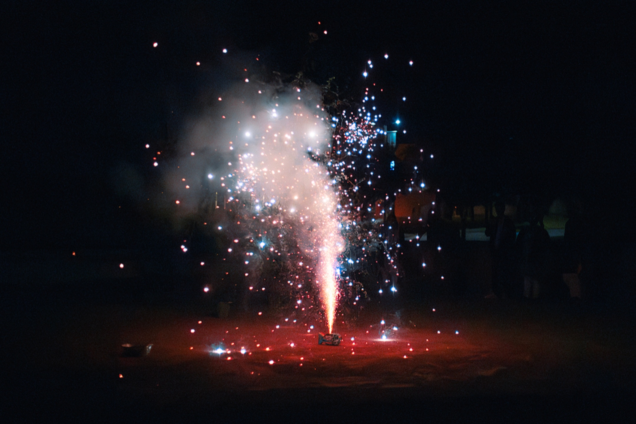 The image shows a vivid, colorful firework display against a dark background. The firework is positioned on the ground and emits a bright, concentrated stream of light vertically upwards. This central beam is a mix of intense red and white colors. Surrounding this, numerous smaller sparks and bursts of light radiate outward in various directions, creating an explosive spectacle of red, blue, and white hues. The scene is enveloped in smoke, enhancing the luminous effects of the firework bursts. In the background, faint silhouettes of people can be seen observing the display from a distance, though the darkness makes them indistinct.