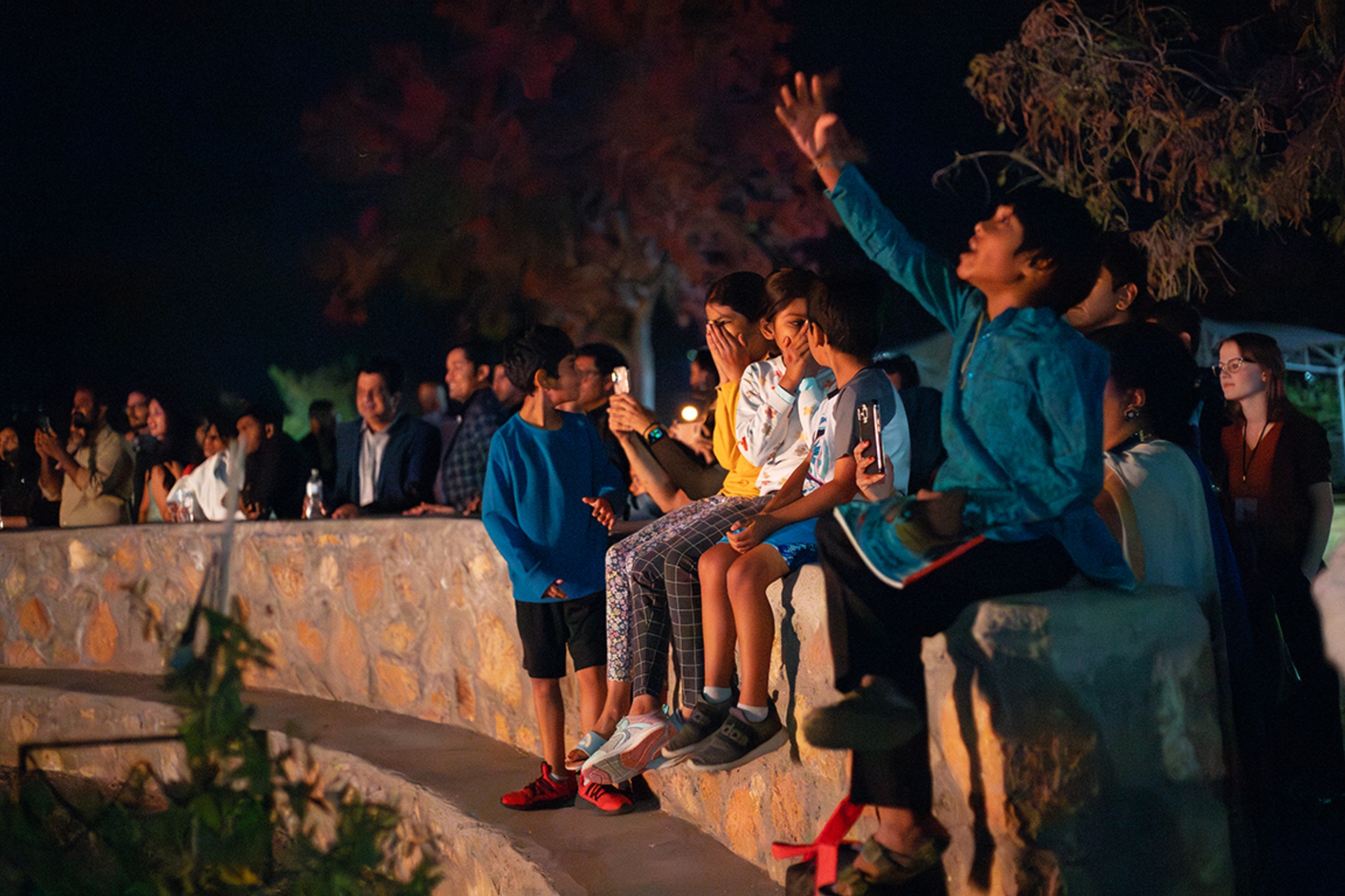 The image portrays a group of people, including children and adults, gathered outdoors at night, likely watching an event or performance. The scene is lit by artificial lights creating a warm glow. The people are standing or sitting on a stone structure composed of light-colored, irregularly shaped stones. The foreground left side shows several leafy branches, hinting at surrounding vegetation. In the background, there are more people standing near a dimly lit area, and some distant trees are softly visible. One child in a blue sweatshirt and red shoes stands on the ground, while others, mostly children, sit on the stone ledge, some holding phones to capture the moment. A child near the right side is raising an arm excitedly, and a woman in a red top stands further in the background on the right, focusing on the event.