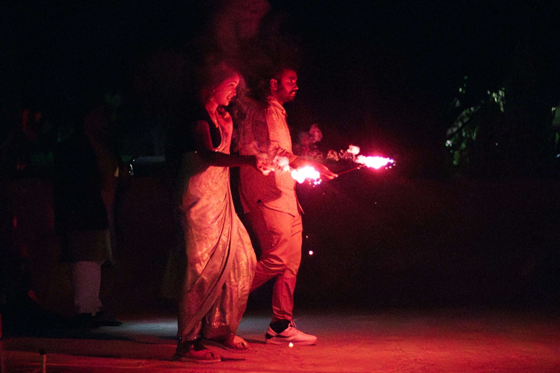 The image captures a nighttime scene where two people are celebrating with sparklers. The woman on the left is wearing a traditional sari, which is illuminated by the bright light of the sparklers in her hand. The man on the right is casually dressed in trousers and a shirt, holding a sparkler of his own. The brightness of the sparklers casts a reddish glow on their faces and clothes, emphasizing their expressions and movements. The background is dark, with only faint hints of other people and surroundings barely visible due to the low light.