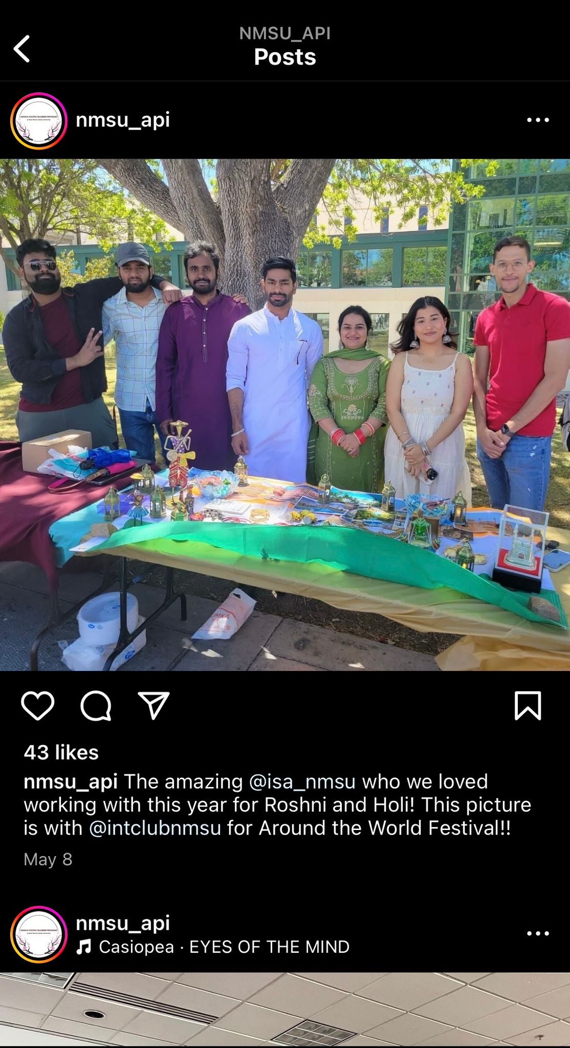 The image portrays a group of seven people standing together outdoors under a large tree with a green and glass building in the background. They appear to be participating in an event, standing behind a table that is decorated with various colorful items, including ornaments, textiles, and small statuettes. The table is draped with a green, yellow, and white cloth. The group is casually dressed in a mix of traditional and modern attire. The overall atmosphere seems celebratory and communal.