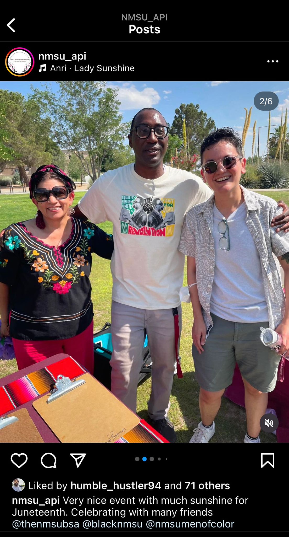 The image shows three people standing close together outdoors in a sunny setting. The person on the left is wearing a black blouse with colorful floral embroidery, large black sunglasses, and has a headband. The person in the middle is taller, wearing glasses, and a white T-shirt with colorful artwork and the words "Equality," "Freedom," "Unite," "Revolution," and "Fight for Justice." The person on the right is wearing a light gray patterned short-sleeve shirt over a white T-shirt, sunglasses, and is holding a water bottle. They are all smiling. The background shows a clear, blue sky with a few clouds, green trees, and a grassy area. In the foreground, there are red tables with clipboards on them.