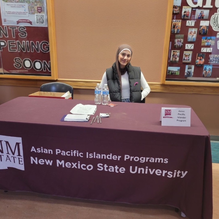 The image shows a promotional setup for New Mexico State University's Asian Pacific Islander Programs. A person is seated at a table covered with a maroon tablecloth that has the university's logo and the program name printed on it. The table has several items including water bottles, pamphlets, and pens. Behind the individual, notice boards display various photos and notices related to university events. The left notice board includes large white letters forming the words "EVENTS HAPPENING SOON."  Alt-text:  Person seated at a New Mexico State University Asian Pacific Islander Programs table with informational materials.  Transcribed Text:  EVENTS HAPPENING SOON Asian Pacific Islander Programs New Mexico State University