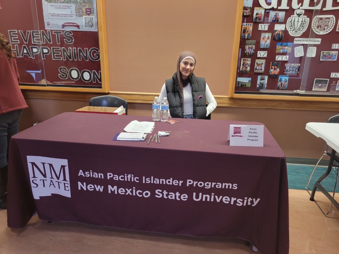 The image shows a promotional setup for New Mexico State University's Asian Pacific Islander Programs. A person is seated at a table covered with a maroon tablecloth that has the university's logo and the program name printed on it. The table has several items including water bottles, pamphlets, and pens. Behind the individual, notice boards display various photos and notices related to university events. The left notice board includes large white letters forming the words "EVENTS HAPPENING SOON."  Alt-text:  Person seated at a New Mexico State University Asian Pacific Islander Programs table with informational materials.  Transcribed Text:  EVENTS HAPPENING SOON Asian Pacific Islander Programs New Mexico State University