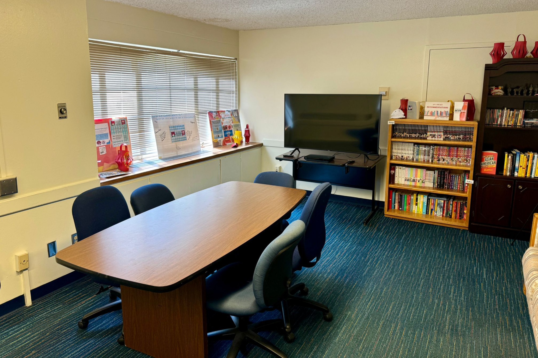 The image depicts a small meeting or study room. The room has blue-striped carpet flooring and off-white walls. A rectangular wooden table with rounded corners is placed centrally, surrounded by six blue swivel chairs. To the left, there is a wide window with horizontal blinds, and the windowsill is adorned with various colorful displays and posters. On the right side, against the wall, there is a large flat-screen TV mounted on a black stand. Next to it, there is a wooden bookshelf filled with a variety of books, including manga and novels. Some decorative red items are on top of the bookshelf and nearby cabinets. The room is well-lit with natural light coming through the window.