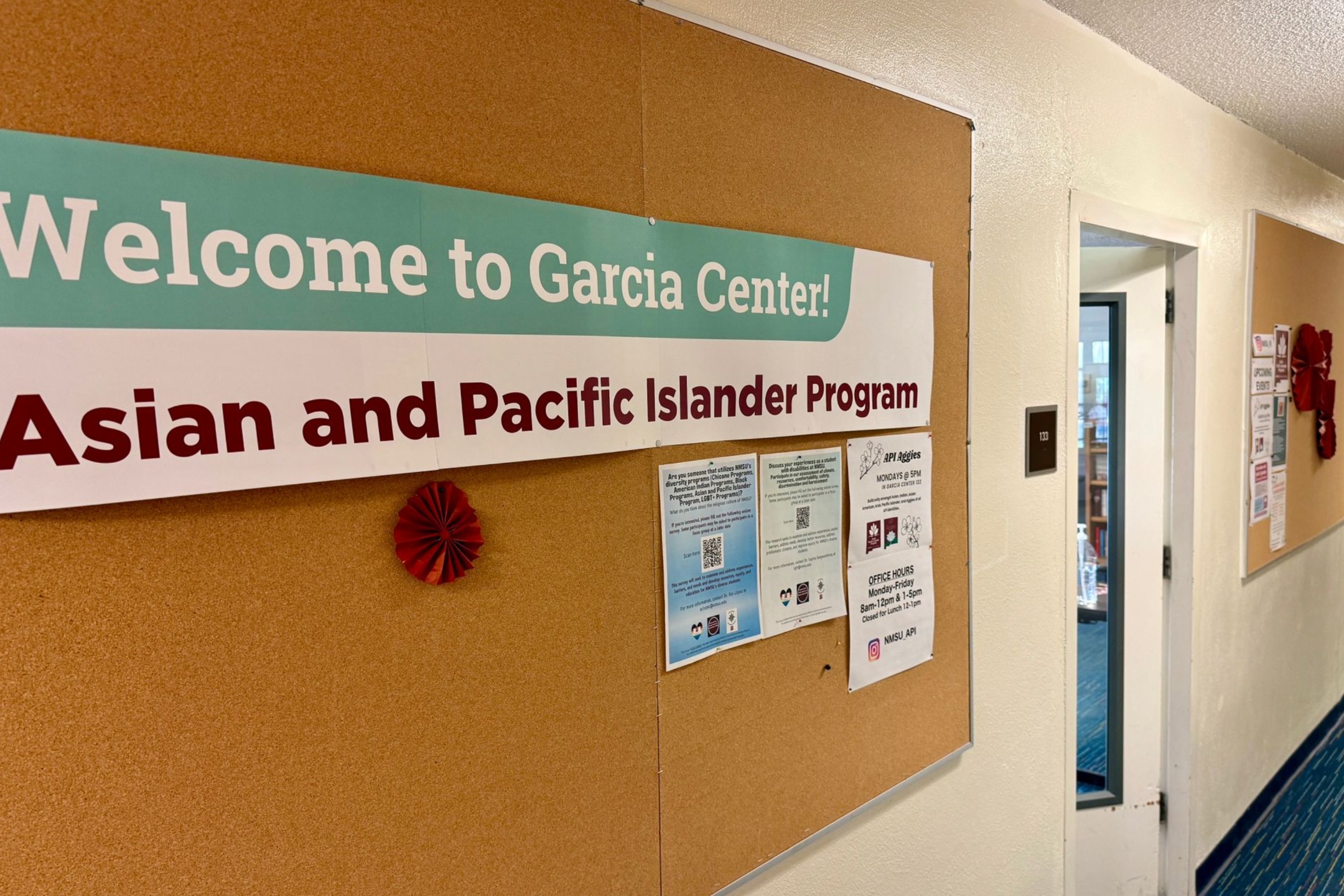  The image shows a corridor with beige walls and blue carpeted flooring. A large cork bulletin board is mounted on the wall to the left. There is a banner on the bulletin board with a mint green and white background, and text in white and maroon. Below the banner are several flyers, each with different information and some featuring QR codes. On the right side of the image, part of an open door reveals a glimpse into another room, and the room number "133" is visible on a sign next to the door. Additionally, there are a couple of decorations on the bulletin board, including a red paper fan.