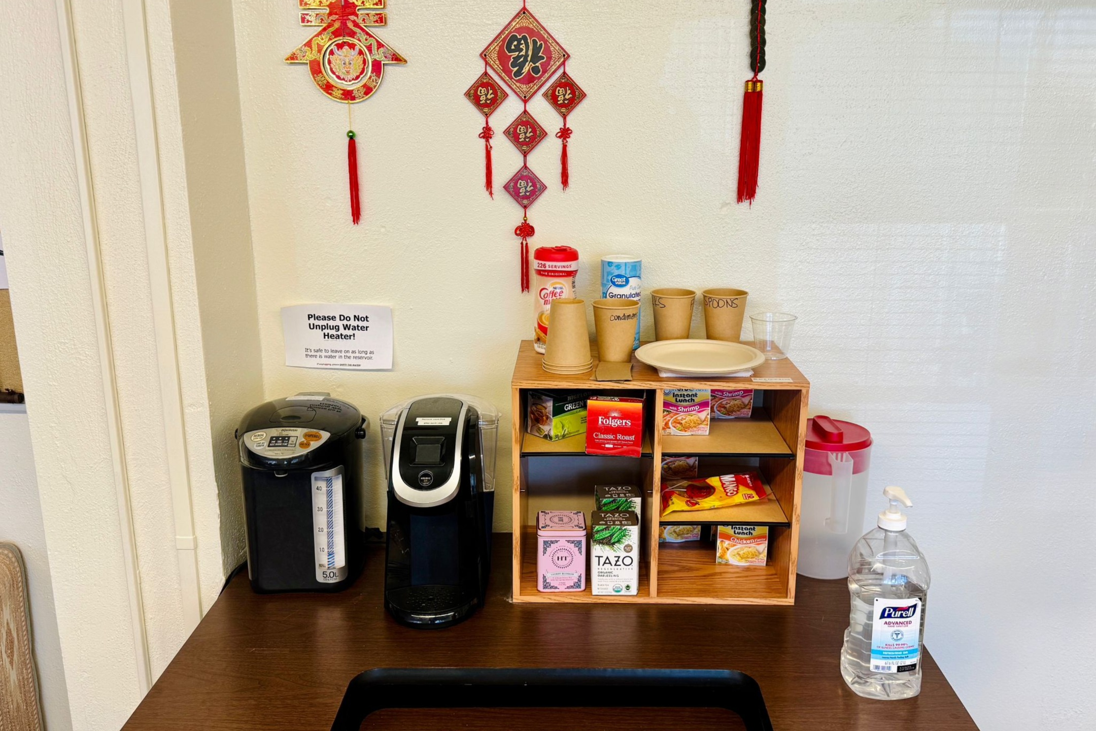 Office refreshment area with a hot water dispenser, Keurig machine, paper cups, tea and coffee supplies, instant noodles, and a bottle of hand sanitizer on a wooden countertop. Red and gold Chinese-style decorations hang on the wall above.