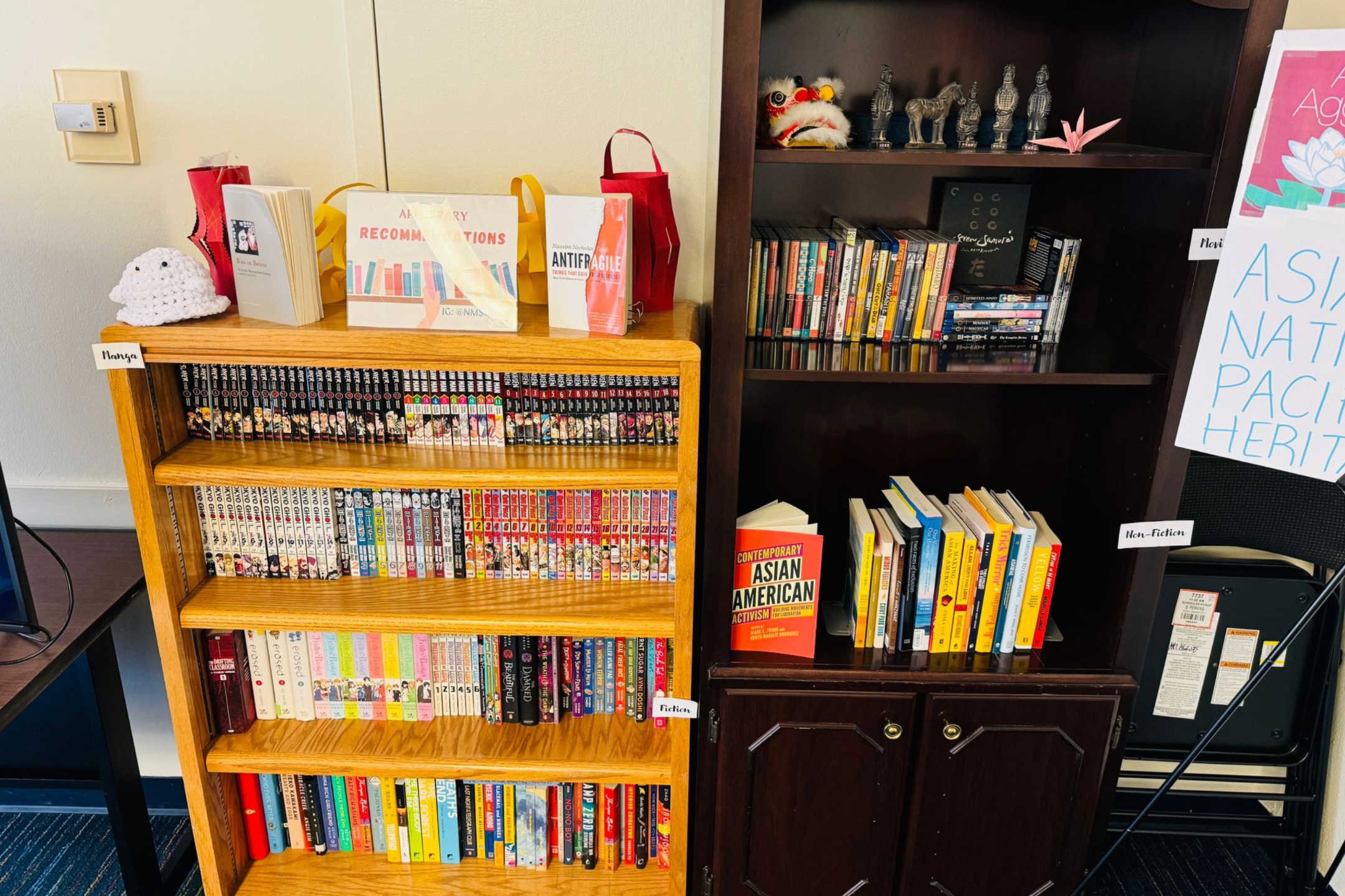  The image showcases two bookshelves placed side by side against a light-colored wall. The left bookshelf, made of light wood, is labeled "Manga" and contains five shelves filled with manga books, characterized by their colorful spines. The top of this bookshelf has decorative items, including a small, white, crocheted toy, two books, and a sign that reads "Art Library Recommendations." The right bookshelf is dark wood and labeled "Non-Fiction." It has three upper shelves filled with books and decorative items, including small figurines, a traditional Chinese dragon head, and an origami crane. The middle shelf holds various books, while the bottom shelf has a mix of books and other items. A book titled "Contemporary Asian American Activism" is prominently displayed standing upright on the bottom shelf. Beside this bookshelf, a poster partially visible reads "Asian N American Pacif Herit." The corner of a desk is visible on the left edge of the image.