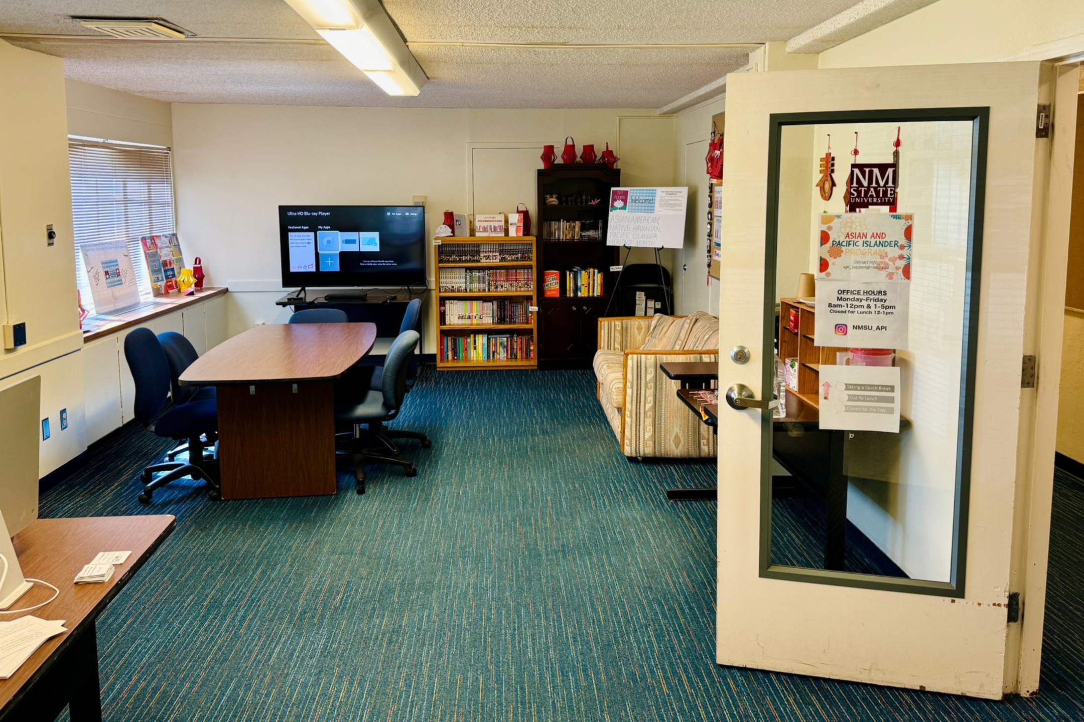 Small room with blue carpet, table with chairs, bookshelves, TV, and a couch. Door poster lists NM State University Asian and Pacific Islander Resource Center office hours.