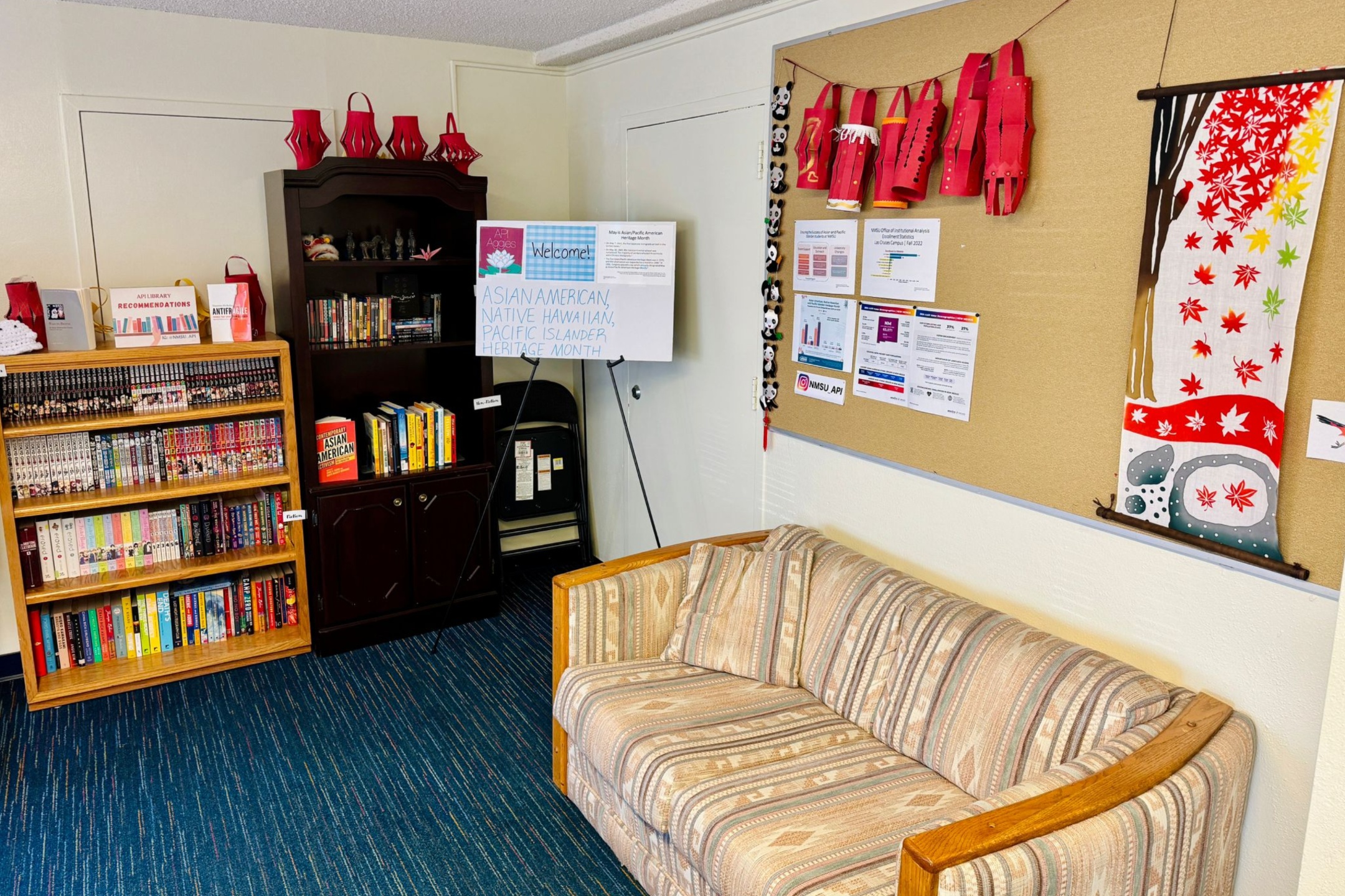 The image depicts a cozy, well-organized room with a library and Asian cultural decorations. To the left, there are two wooden bookshelves filled with books. The top shelf of the leftmost bookshelf contains magazines and the shelves below hold manga and colorful paperbacks. The bookshelf to the right has a mix of books and some ornamental items. Above this bookshelf are red paper lanterns. In the center of the image, there is a display board on an easel with a welcome sign for Asian American, Native Hawaiian, and Pacific Islander Heritage Month. Next to the display board, there are two folded black chairs leaning against a white door. The right side of the room features a large corkboard filled with various flyers, a string of panda plushies, and red paper lanterns. Below the corkboard, there is a beige, patterned sofa with two cushions.