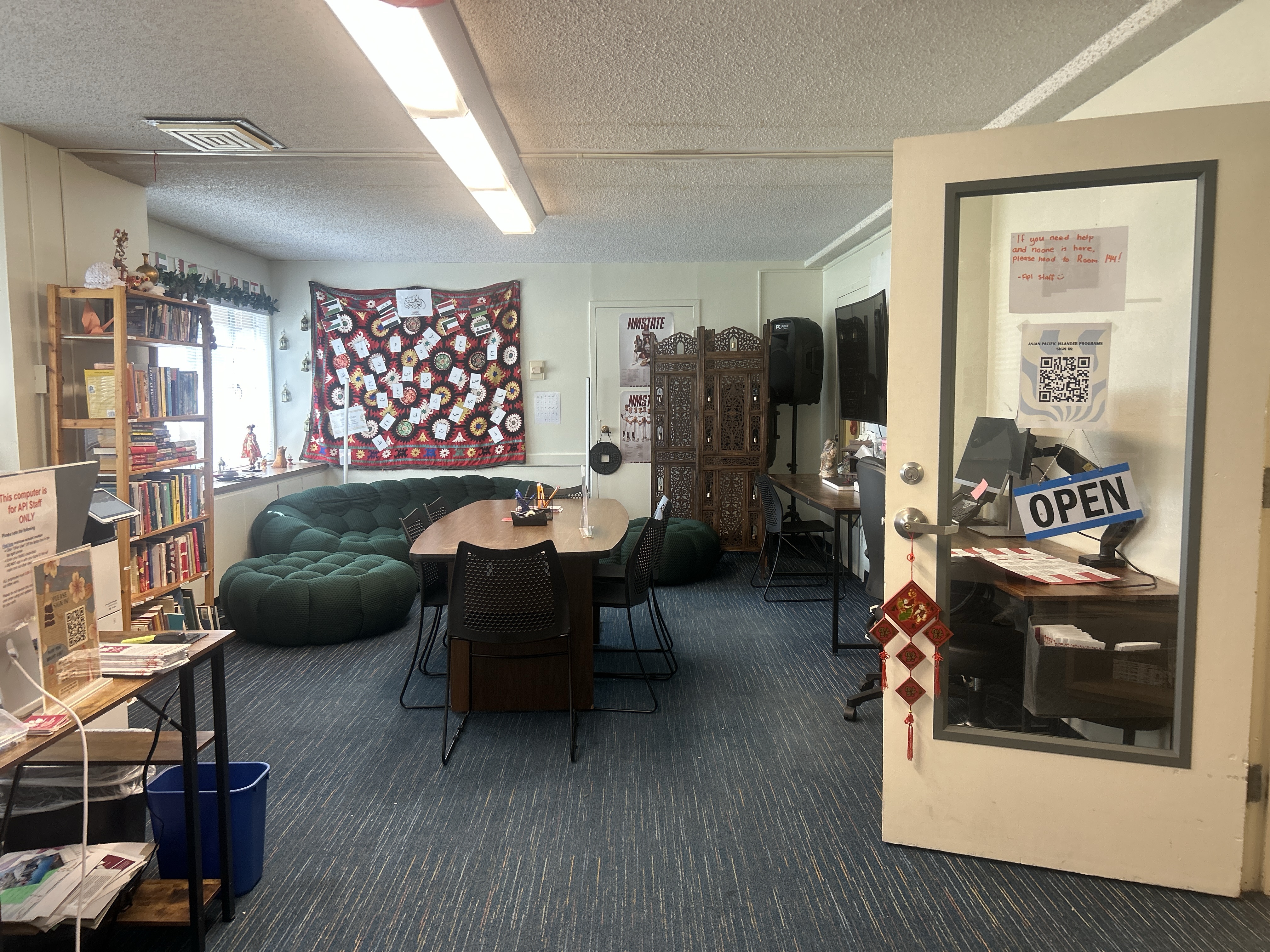 Small room with blue carpet, table with chairs, bookshelves, TV, and a couch. Door poster lists NM State University Asian and Pacific Islander Resource Center office hours.