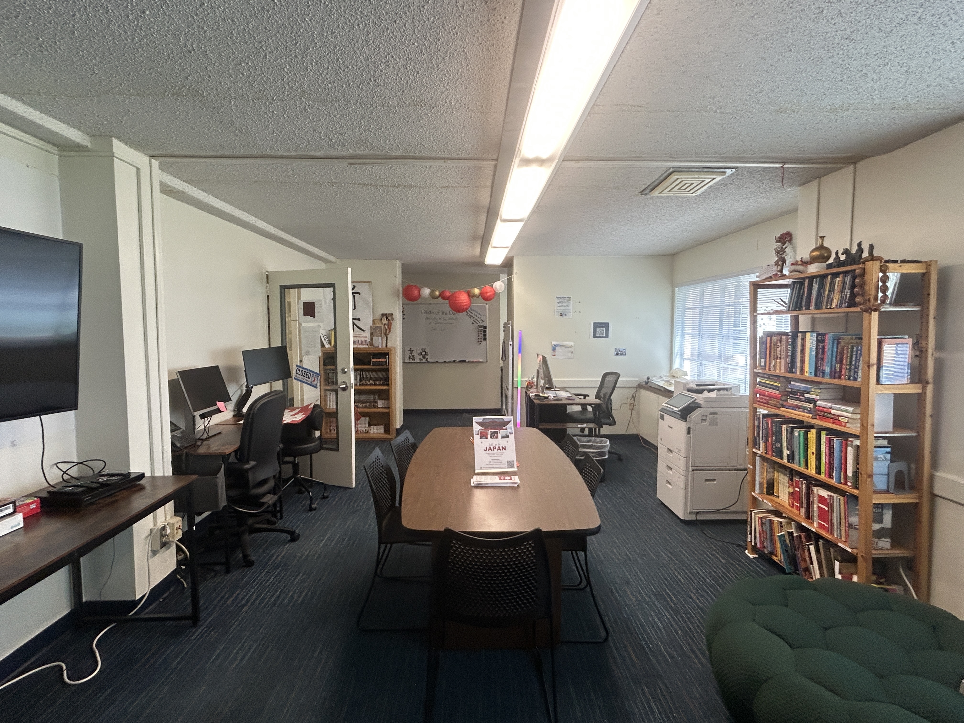 The image displays an office space with a desk centered in the foreground. The desk is light brown with black trim and is arranged facing towards a wall on the right side of the image. On the desk, there is an Apple iMac computer, a keyboard, a wired mouse, a black office phone, and various office supplies such as a container of hand sanitizer, a pen holder, and sticky notes. The chair at the desk has a light grey fabric seat and black armrests.  On the wall to the right of the desk, there is a sign which reads, “THIS AREA, CHAIR, & COMPUTER ARE RESERVED FOR API PROGRAM STAFF ONLY.” Additional papers and a small chart are also affixed to this wall.  In the background, to the left, there is an open doorway leading to another room. The wall adjacent to this doorway is decorated with multiple printed sheets of paper and photographs, showcasing what appears to be content related to "Asian and Pacific Islander History." Below these papers, a broom and a dustpan are placed beside a black trash bin.  The flooring is blue carpet with green and yellow stripes running through it, complementing the clean and structured configuration of the entire office space.
