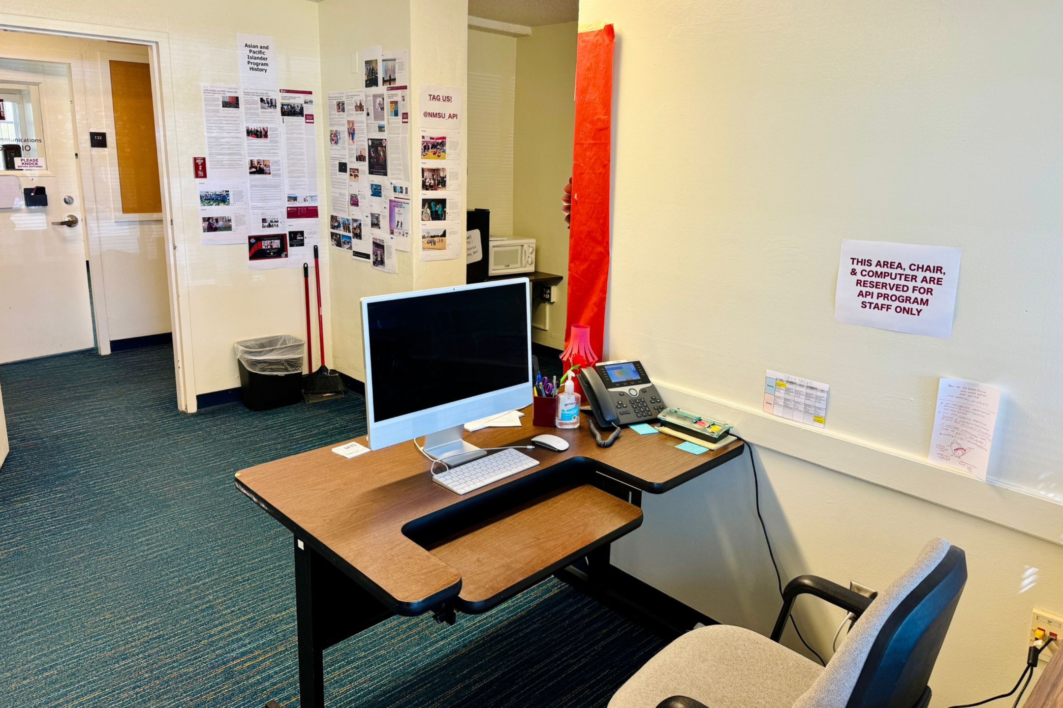 The image displays an office space with a desk centered in the foreground. The desk is light brown with black trim and is arranged facing towards a wall on the right side of the image. On the desk, there is an Apple iMac computer, a keyboard, a wired mouse, a black office phone, and various office supplies such as a container of hand sanitizer, a pen holder, and sticky notes. The chair at the desk has a light grey fabric seat and black armrests.  On the wall to the right of the desk, there is a sign which reads, “THIS AREA, CHAIR, & COMPUTER ARE RESERVED FOR API PROGRAM STAFF ONLY.” Additional papers and a small chart are also affixed to this wall.  In the background, to the left, there is an open doorway leading to another room. The wall adjacent to this doorway is decorated with multiple printed sheets of paper and photographs, showcasing what appears to be content related to "Asian and Pacific Islander History." Below these papers, a broom and a dustpan are placed beside a black trash bin.  The flooring is blue carpet with green and yellow stripes running through it, complementing the clean and structured configuration of the entire office space.