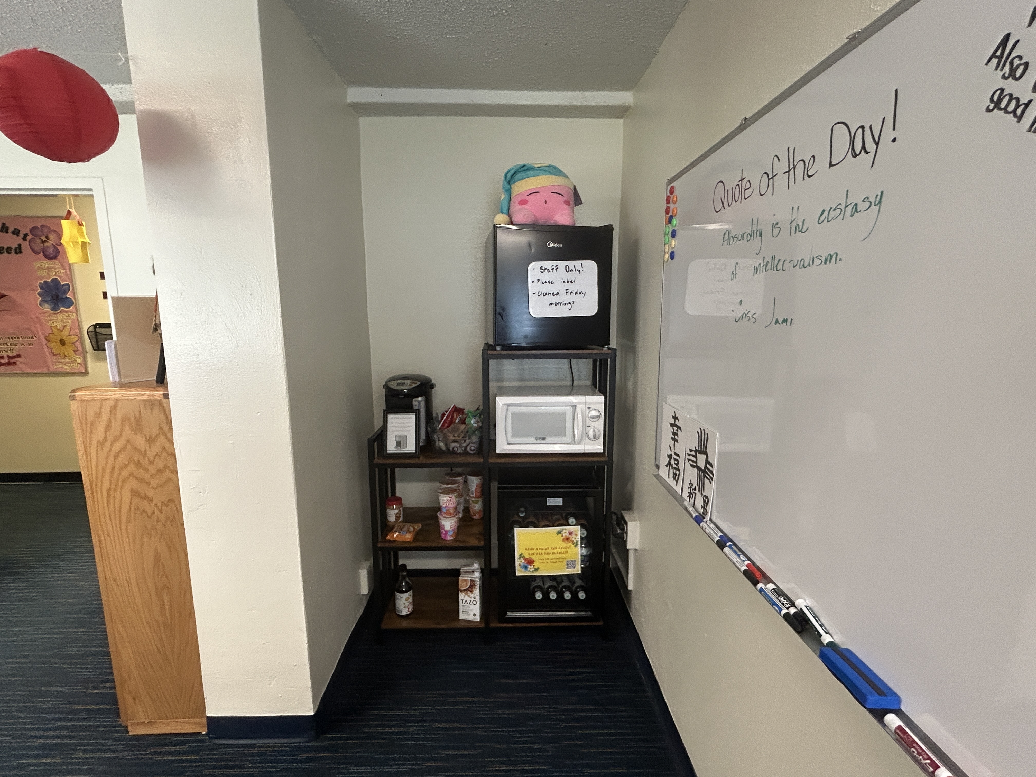 The image shows a row of three desktop computers on a wooden table. Each computer consists of a black monitor labeled "SCEPTRE" on the bottom-left corner, a black keyboard, and a black mouse. The monitors are placed on top of black CPU units. Behind the table, there is a large window with horizontal blinds that are partially closed, allowing some daylight to filter through. In front of each computer setup is an office chair, two of which are cushioned with black seats and one with fabric. To the right of the table and computers, there is a wooden bookshelf containing several pieces of red decorative items on display.