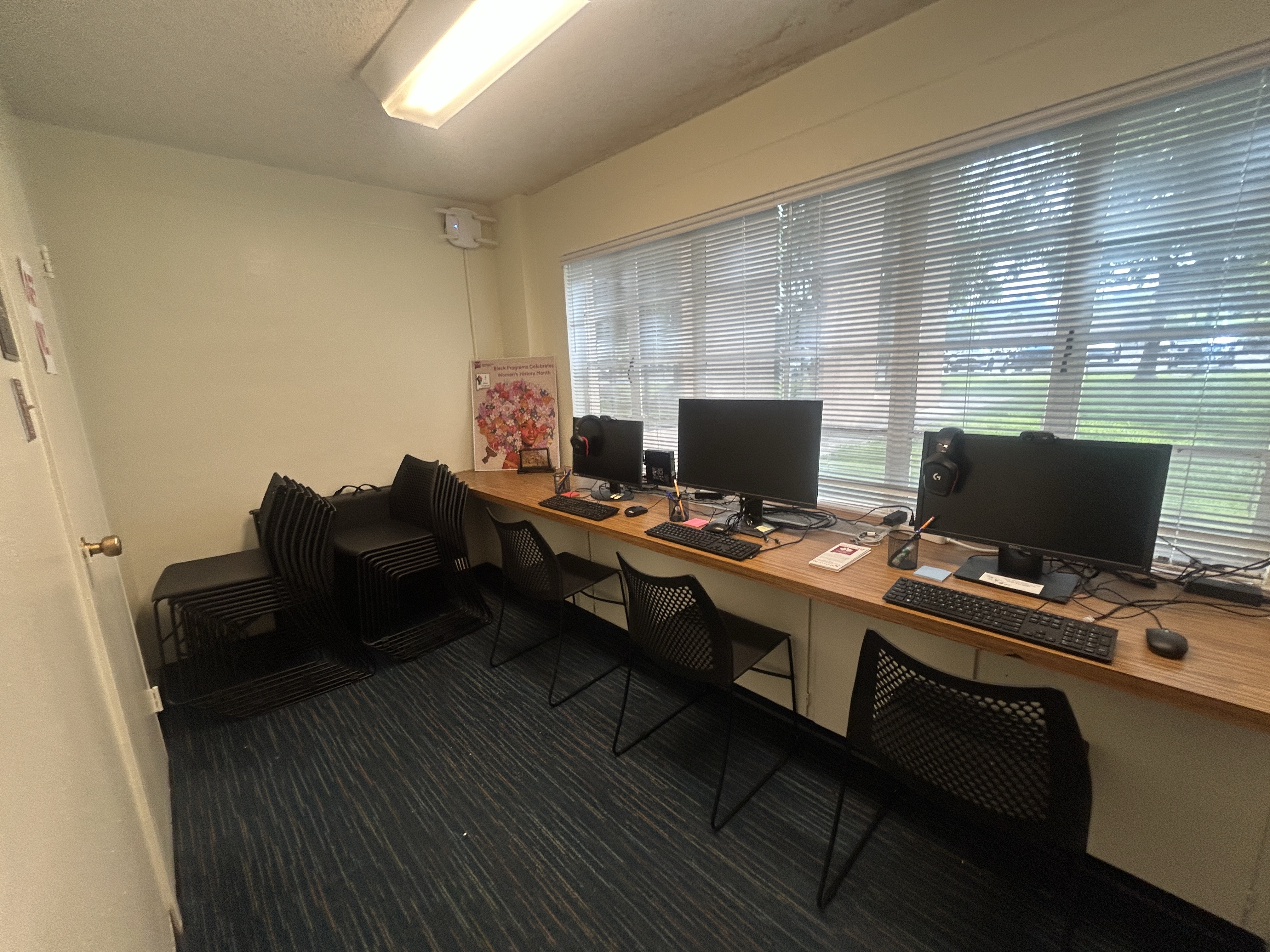 Office refreshment area with a hot water dispenser, Keurig machine, paper cups, tea and coffee supplies, instant noodles, and a bottle of hand sanitizer on a wooden countertop. Red and gold Chinese-style decorations hang on the wall above.
