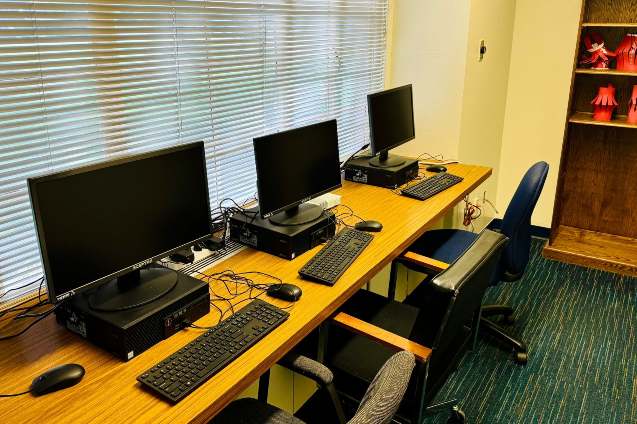 The image shows a row of three desktop computers on a wooden table. Each computer consists of a black monitor labeled "SCEPTRE" on the bottom-left corner, a black keyboard, and a black mouse. The monitors are placed on top of black CPU units. Behind the table, there is a large window with horizontal blinds that are partially closed, allowing some daylight to filter through. In front of each computer setup is an office chair, two of which are cushioned with black seats and one with fabric. To the right of the table and computers, there is a wooden bookshelf containing several pieces of red decorative items on display.