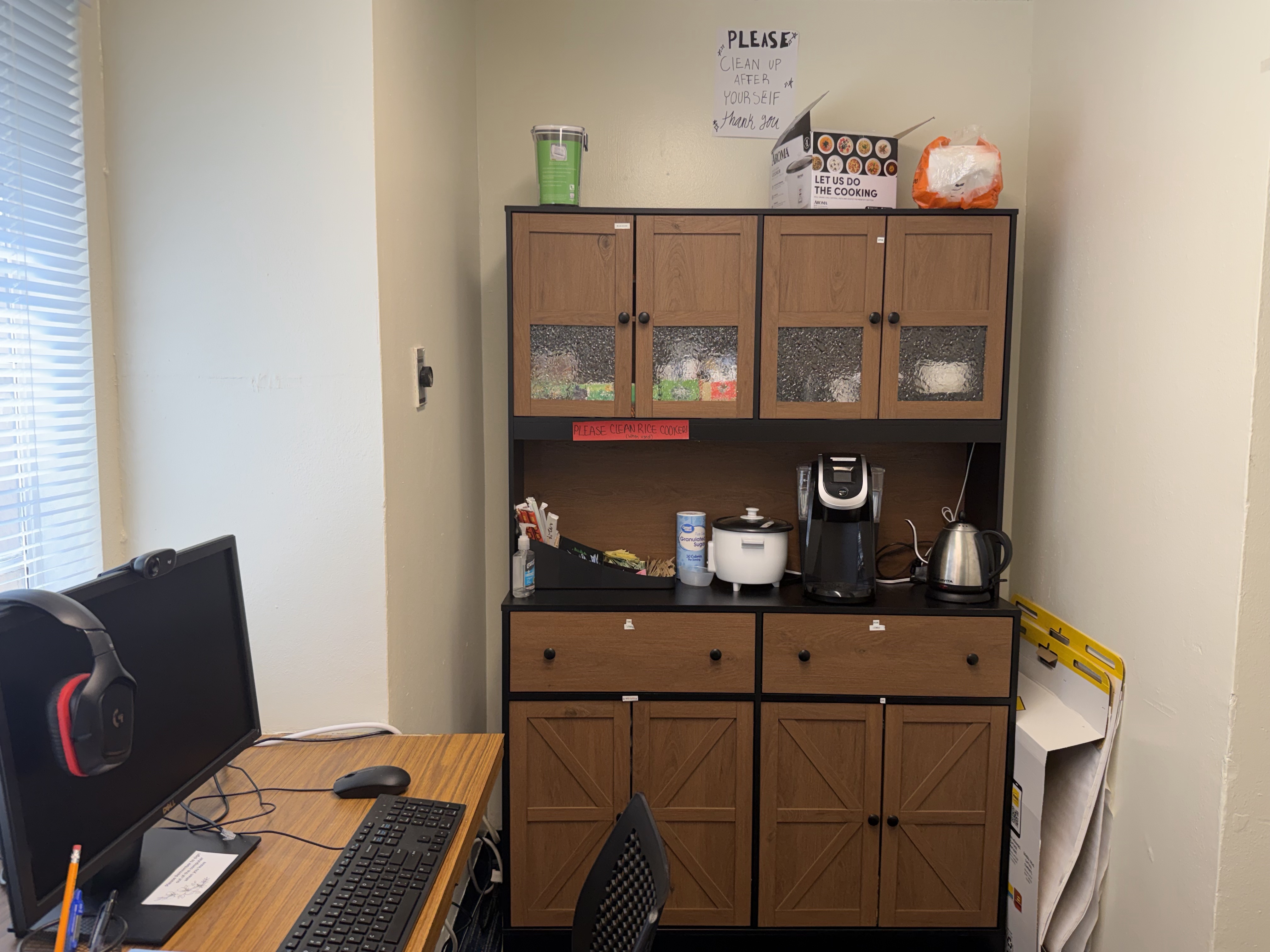 The image depicts a small meeting or study room. The room has blue-striped carpet flooring and off-white walls. A rectangular wooden table with rounded corners is placed centrally, surrounded by six blue swivel chairs. To the left, there is a wide window with horizontal blinds, and the windowsill is adorned with various colorful displays and posters. On the right side, against the wall, there is a large flat-screen TV mounted on a black stand. Next to it, there is a wooden bookshelf filled with a variety of books, including manga and novels. Some decorative red items are on top of the bookshelf and nearby cabinets. The room is well-lit with natural light coming through the window.